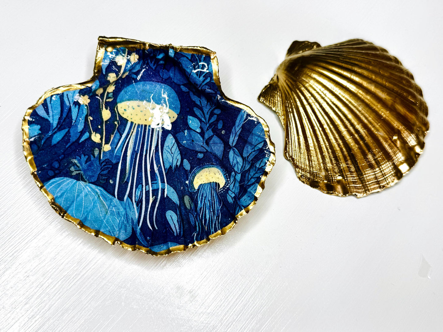 Hand-gilded oyster shell and scallop shell dishes featuring an elegant blue jellyfish design with ocean botanicals, finished with real liquid gold leaf and crystal-clear resin. Coastal ring dishes and jewellery holders available individually or as a matching shell gift set. ✨