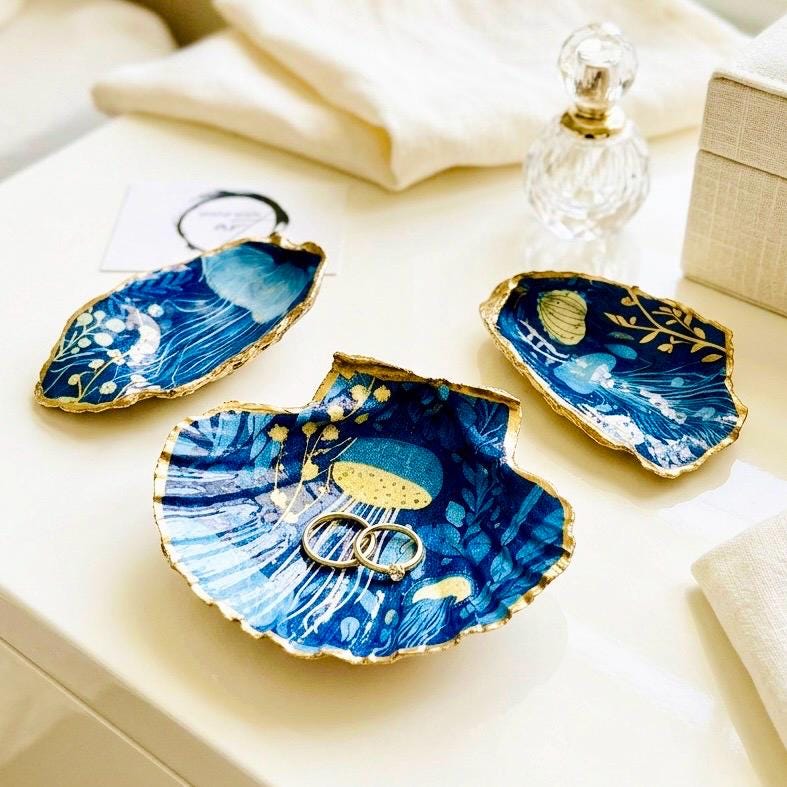 Hand-gilded oyster shell and scallop shell dishes featuring an elegant blue jellyfish design with ocean botanicals, finished with real liquid gold leaf and crystal-clear resin. Coastal ring dishes and jewellery holders available individually or as a matching shell gift set. ✨