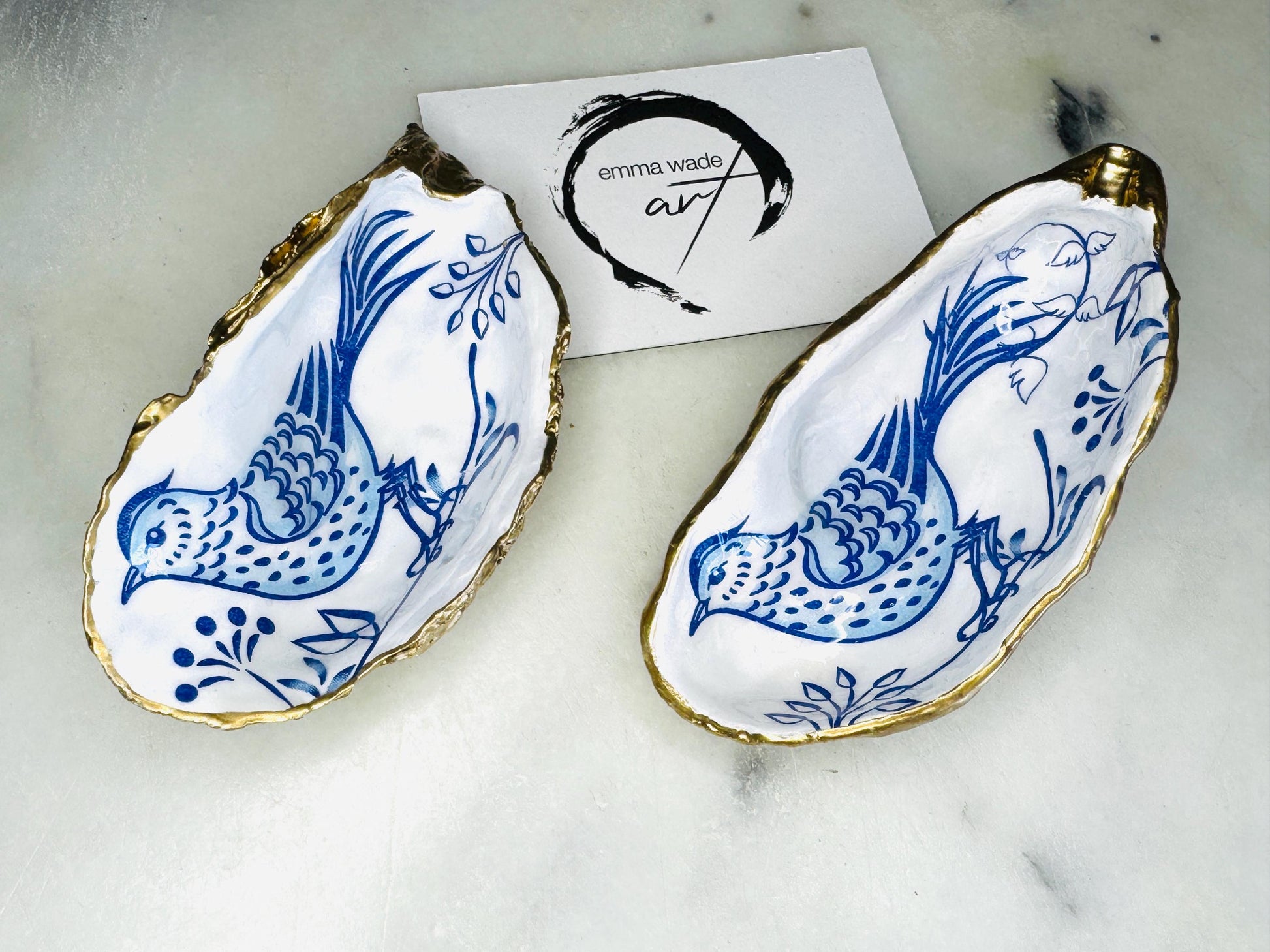 Handmade blue bird oyster shell salt and pepper pinch pot set crafted from natural Dorset oyster shells with glossy pearlescent interiors, blue porcelain style bird artwork and shimmering liquid gold leaf edging. Luxury coastal cruet dishes sealed with gloss resin and styled with optional gold spoons. Elegant seaside dining decor for beach house kitchens, dinner parties, weddings, anniversaries and bird lovers. Artisan shell tableware handmade in the UK with coastal blue and white styling.