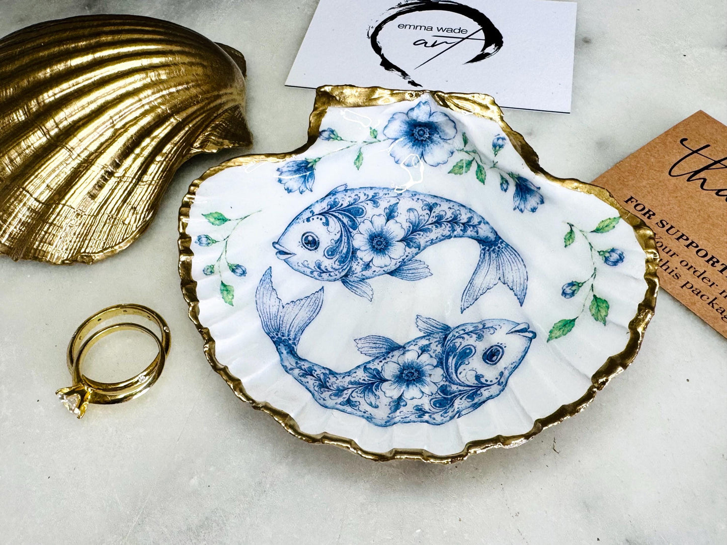 Handcrafted scallop shell trinket dish featuring an original Delft Blue fish illustration created and designed by Emma Wade ART. Natural scallop shell finished with a choice of genuine gold leaf or silver leaf and sealed beneath crystal-hard glossy resin. Coastal ring holder, jewellery dish and decorative shell tray inspired by traditional Delft ceramics and marine life. Available in standard 10–11cm or large 12–14cm sizes. Part of the Delft Blue Sea Life Collection.
