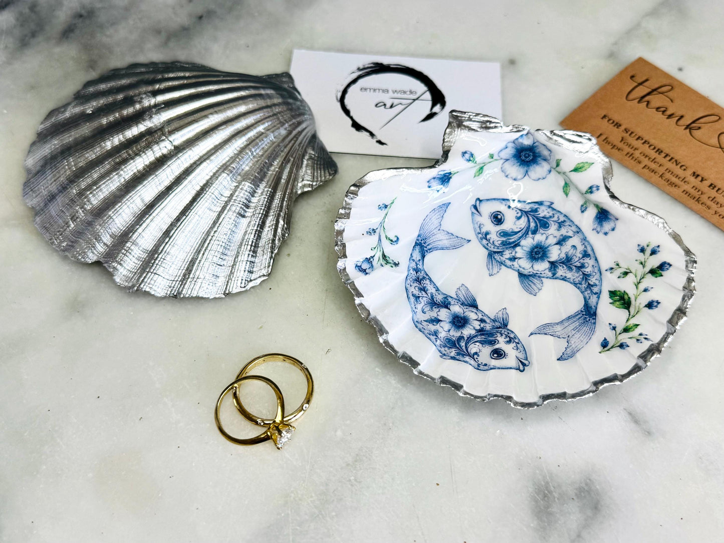 Handcrafted scallop shell trinket dish featuring an original Delft Blue fish illustration created and designed by Emma Wade ART. Natural scallop shell finished with a choice of genuine gold leaf or silver leaf and sealed beneath crystal-hard glossy resin. Coastal ring holder, jewellery dish and decorative shell tray inspired by traditional Delft ceramics and marine life. Available in standard 10–11cm or large 12–14cm sizes. Part of the Delft Blue Sea Life Collection.