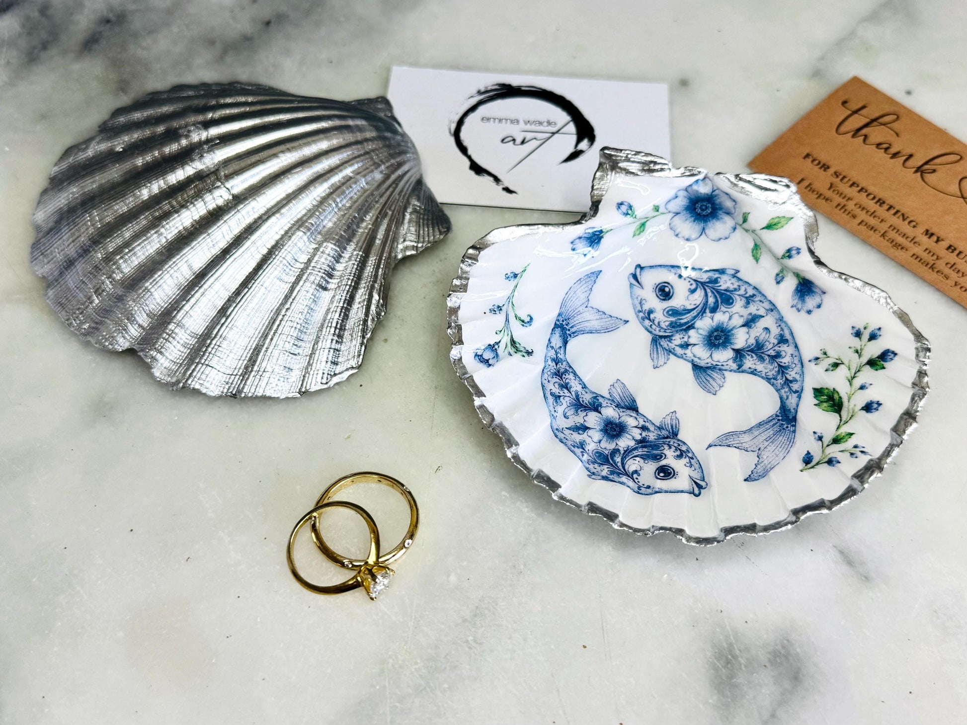 Handcrafted scallop shell trinket dish featuring an original Delft Blue fish illustration created and designed by Emma Wade ART. Natural scallop shell finished with a choice of genuine gold leaf or silver leaf and sealed beneath crystal-hard glossy resin. Coastal ring holder, jewellery dish and decorative shell tray inspired by traditional Delft ceramics and marine life. Available in standard 10–11cm or large 12–14cm sizes. Part of the Delft Blue Sea Life Collection.