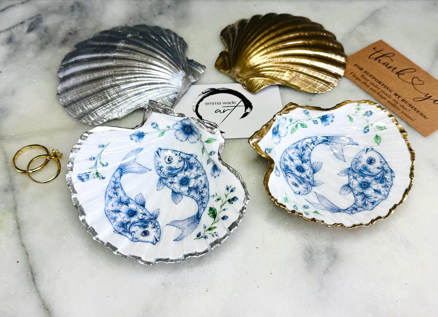 Handcrafted scallop shell trinket dish featuring an original Delft Blue fish illustration created and designed by Emma Wade ART. Natural scallop shell finished with a choice of genuine gold leaf or silver leaf and sealed beneath crystal-hard glossy resin. Coastal ring holder, jewellery dish and decorative shell tray inspired by traditional Delft ceramics and marine life. Available in standard 10–11cm or large 12–14cm sizes. Part of the Delft Blue Sea Life Collection.
