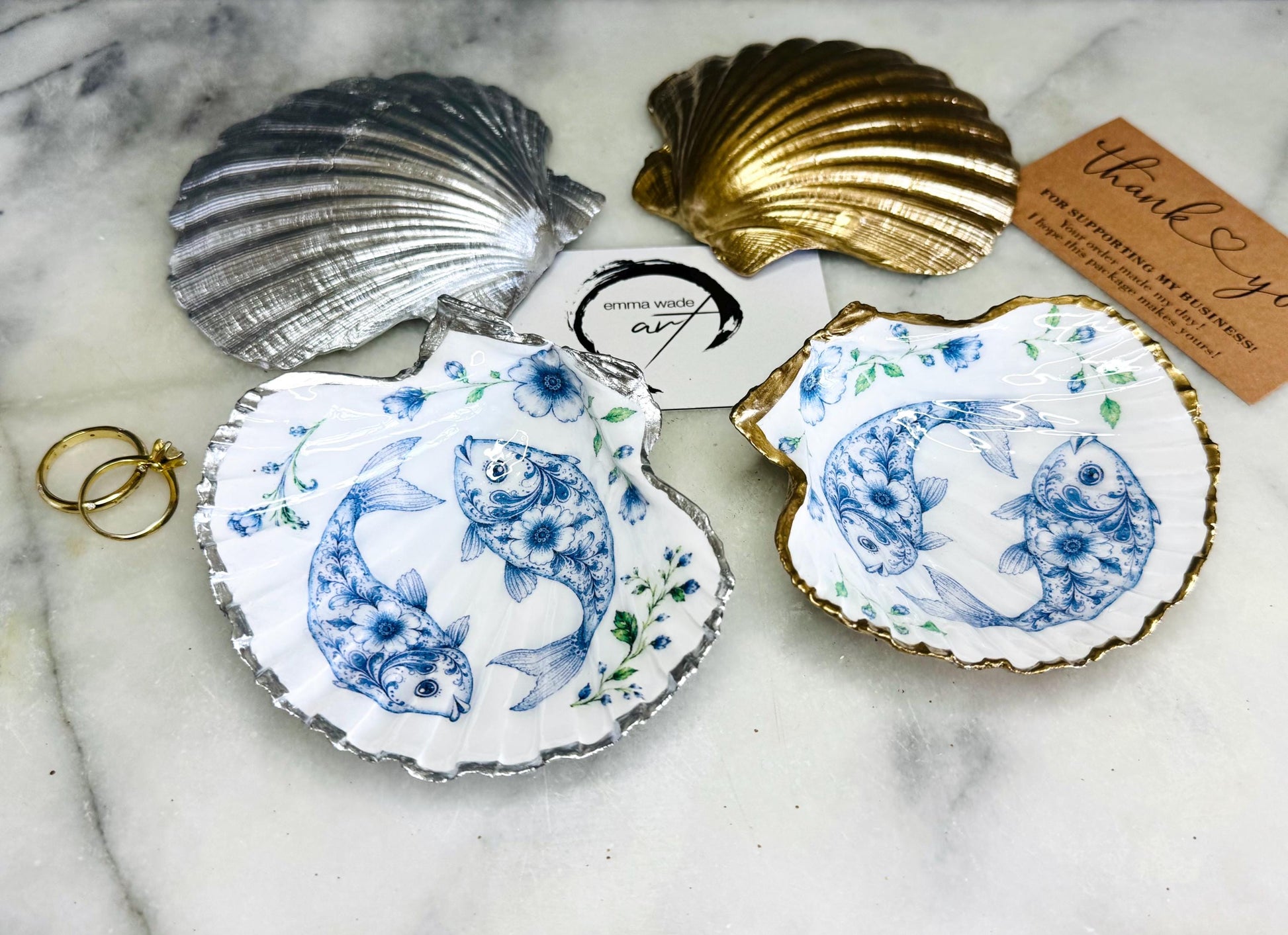 Handcrafted scallop shell trinket dish featuring an original Delft Blue fish illustration created and designed by Emma Wade ART. Natural scallop shell finished with a choice of genuine gold leaf or silver leaf and sealed beneath crystal-hard glossy resin. Coastal ring holder, jewellery dish and decorative shell tray inspired by traditional Delft ceramics and marine life. Available in standard 10–11cm or large 12–14cm sizes. Part of the Delft Blue Sea Life Collection.
