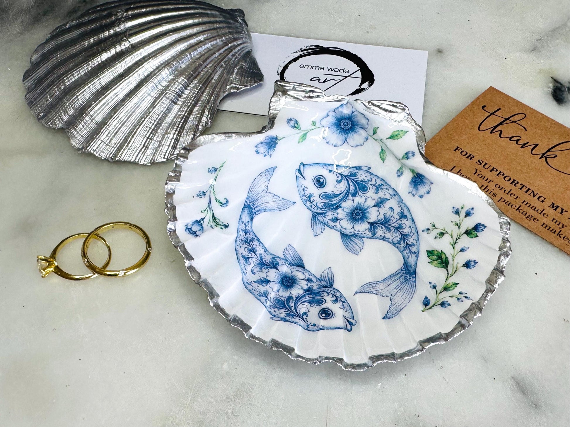 Handcrafted scallop shell trinket dish featuring an original Delft Blue fish illustration created and designed by Emma Wade ART. Natural scallop shell finished with a choice of genuine gold leaf or silver leaf and sealed beneath crystal-hard glossy resin. Coastal ring holder, jewellery dish and decorative shell tray inspired by traditional Delft ceramics and marine life. Available in standard 10–11cm or large 12–14cm sizes. Part of the Delft Blue Sea Life Collection.