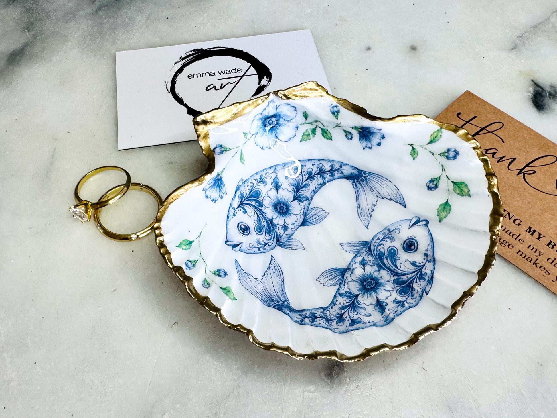 Handcrafted scallop shell trinket dish featuring an original Delft Blue fish illustration created and designed by Emma Wade ART. Natural scallop shell finished with a choice of genuine gold leaf or silver leaf and sealed beneath crystal-hard glossy resin. Coastal ring holder, jewellery dish and decorative shell tray inspired by traditional Delft ceramics and marine life. Available in standard 10–11cm or large 12–14cm sizes. Part of the Delft Blue Sea Life Collection.