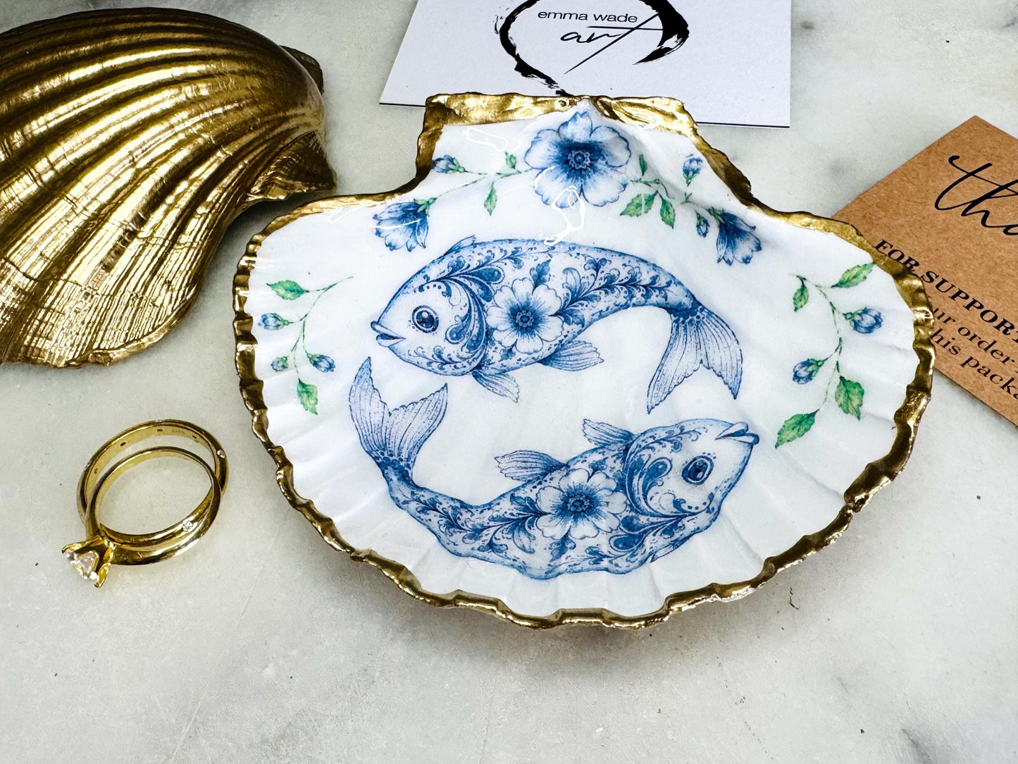 Handcrafted scallop shell trinket dish featuring an original Delft Blue fish illustration created and designed by Emma Wade ART. Natural scallop shell finished with a choice of genuine gold leaf or silver leaf and sealed beneath crystal-hard glossy resin. Coastal ring holder, jewellery dish and decorative shell tray inspired by traditional Delft ceramics and marine life. Available in standard 10–11cm or large 12–14cm sizes. Part of the Delft Blue Sea Life Collection.