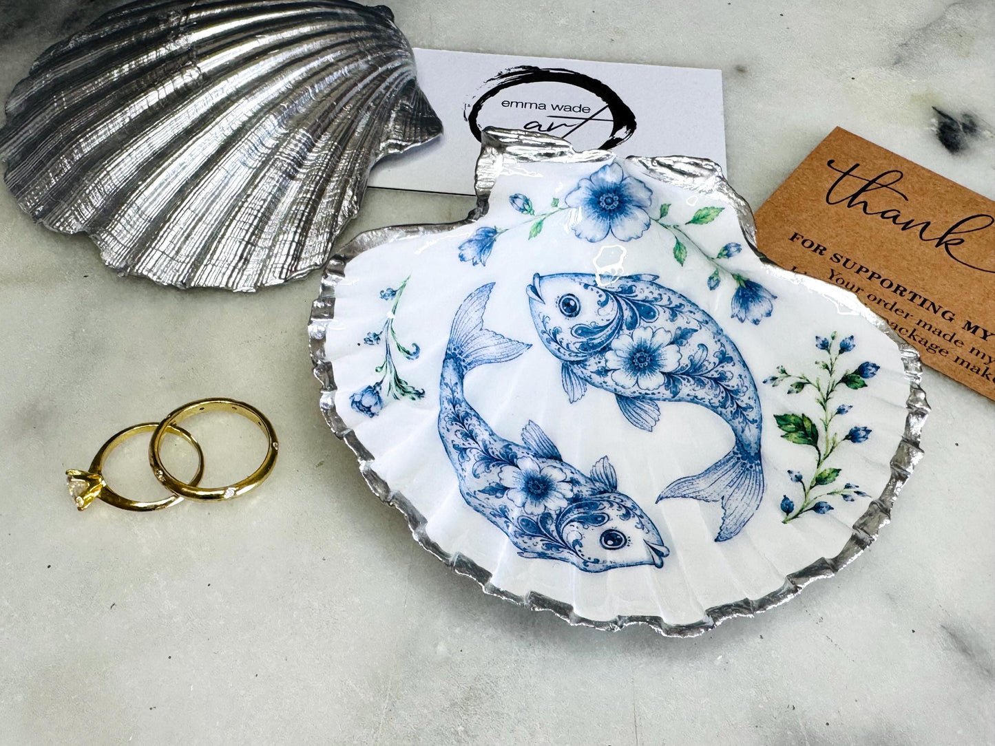 Handcrafted scallop shell trinket dish featuring an original Delft Blue fish illustration created and designed by Emma Wade ART. Natural scallop shell finished with a choice of genuine gold leaf or silver leaf and sealed beneath crystal-hard glossy resin. Coastal ring holder, jewellery dish and decorative shell tray inspired by traditional Delft ceramics and marine life. Available in standard 10–11cm or large 12–14cm sizes. Part of the Delft Blue Sea Life Collection.