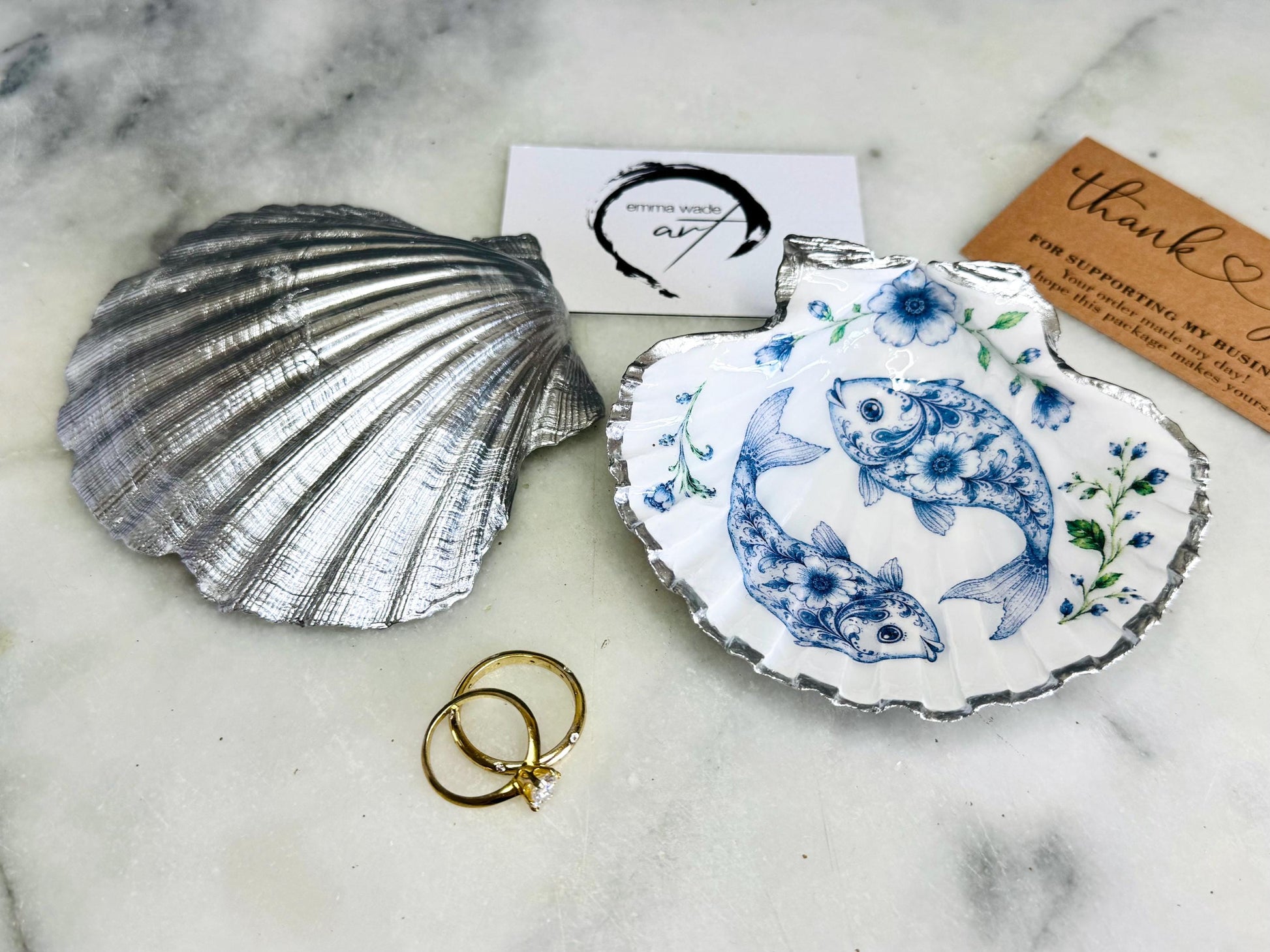 Handcrafted scallop shell trinket dish featuring an original Delft Blue fish illustration created and designed by Emma Wade ART. Natural scallop shell finished with a choice of genuine gold leaf or silver leaf and sealed beneath crystal-hard glossy resin. Coastal ring holder, jewellery dish and decorative shell tray inspired by traditional Delft ceramics and marine life. Available in standard 10–11cm or large 12–14cm sizes. Part of the Delft Blue Sea Life Collection.