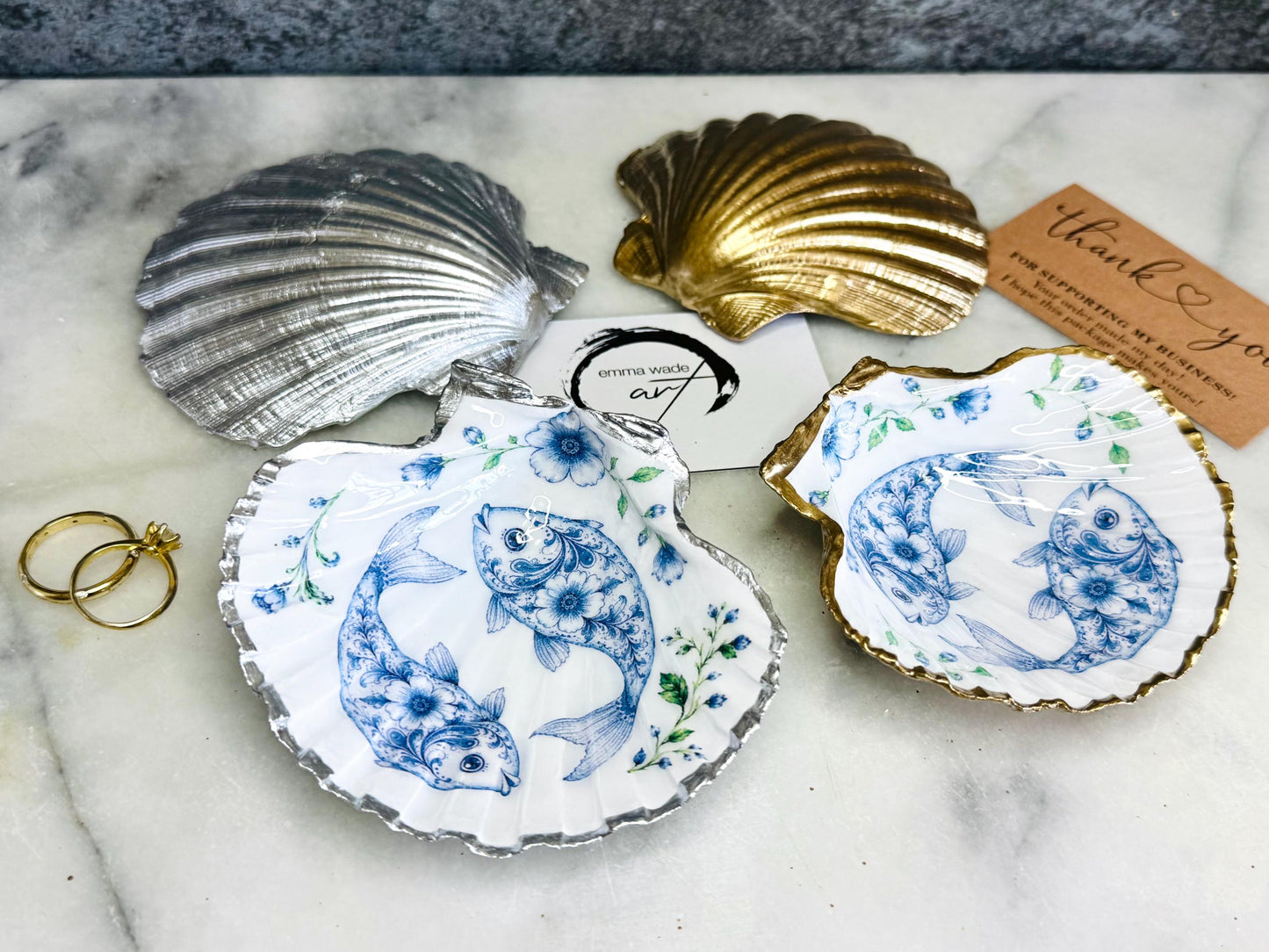 Handcrafted scallop shell trinket dish featuring an original Delft Blue fish illustration created and designed by Emma Wade ART. Natural scallop shell finished with a choice of genuine gold leaf or silver leaf and sealed beneath crystal-hard glossy resin. Coastal ring holder, jewellery dish and decorative shell tray inspired by traditional Delft ceramics and marine life. Available in standard 10–11cm or large 12–14cm sizes. Part of the Delft Blue Sea Life Collection.