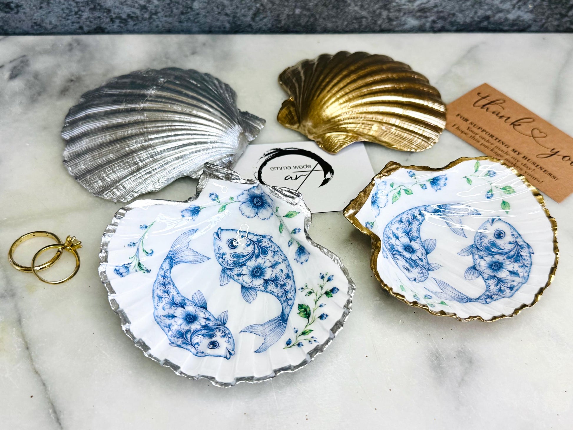 Handcrafted scallop shell trinket dish featuring an original Delft Blue fish illustration created and designed by Emma Wade ART. Natural scallop shell finished with a choice of genuine gold leaf or silver leaf and sealed beneath crystal-hard glossy resin. Coastal ring holder, jewellery dish and decorative shell tray inspired by traditional Delft ceramics and marine life. Available in standard 10–11cm or large 12–14cm sizes. Part of the Delft Blue Sea Life Collection.