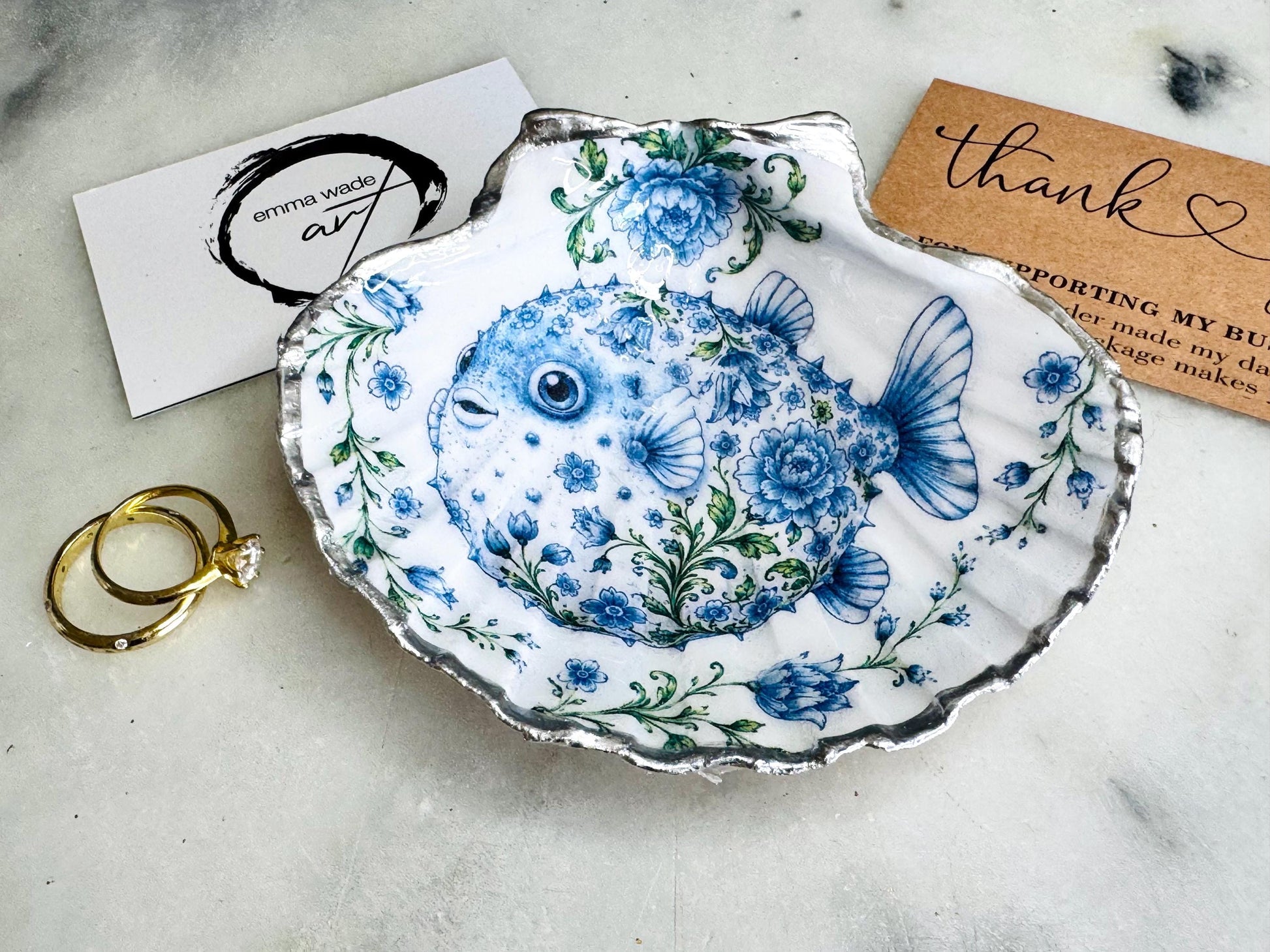 Handcrafted scallop shell trinket dish featuring an original Delft Blue puffer fish illustration created and designed by Emma Wade ART. Natural scallop shell finished with a choice of genuine gold leaf or silver leaf and sealed beneath crystal-hard glossy resin. Coastal ring holder, jewellery dish and decorative shell tray inspired by traditional Delft ceramics and marine life. Available in standard 10–11cm or large 12–14cm sizes.