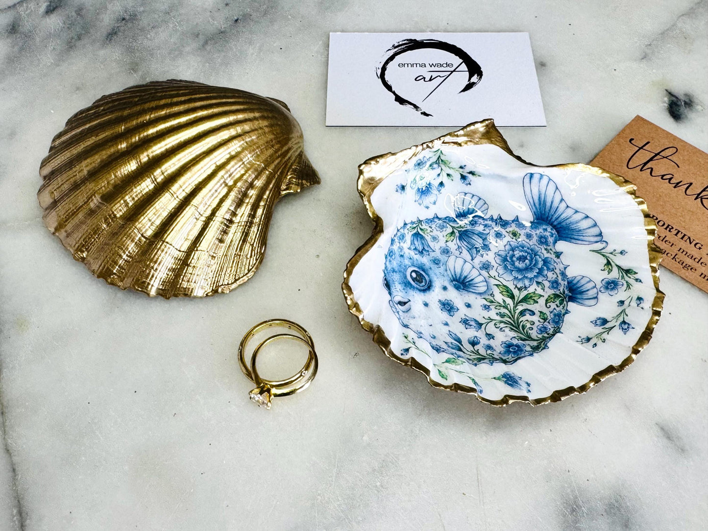 Handcrafted scallop shell trinket dish featuring an original Delft Blue puffer fish illustration created and designed by Emma Wade ART. Natural scallop shell finished with a choice of genuine gold leaf or silver leaf and sealed beneath crystal-hard glossy resin. Coastal ring holder, jewellery dish and decorative shell tray inspired by traditional Delft ceramics and marine life. Available in standard 10–11cm or large 12–14cm sizes.