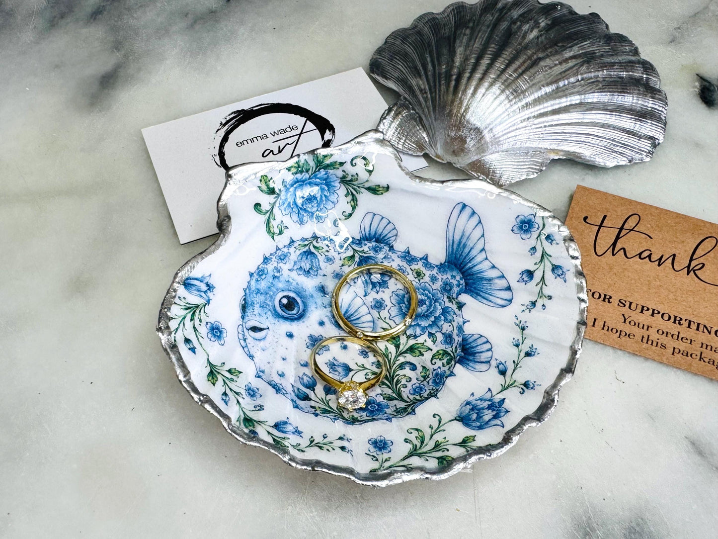 Handcrafted scallop shell trinket dish featuring an original Delft Blue puffer fish illustration created and designed by Emma Wade ART. Natural scallop shell finished with a choice of genuine gold leaf or silver leaf and sealed beneath crystal-hard glossy resin. Coastal ring holder, jewellery dish and decorative shell tray inspired by traditional Delft ceramics and marine life. Available in standard 10–11cm or large 12–14cm sizes.