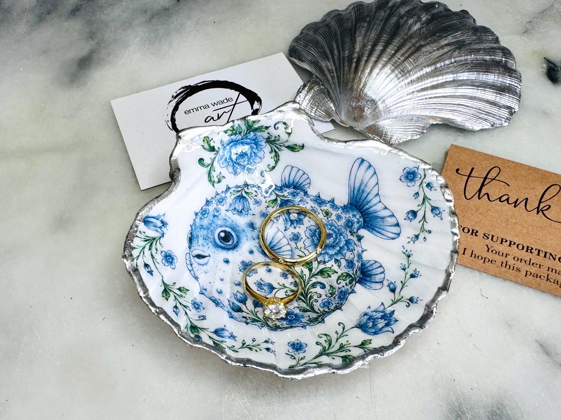 Handcrafted scallop shell trinket dish featuring an original Delft Blue puffer fish illustration created and designed by Emma Wade ART. Natural scallop shell finished with a choice of genuine gold leaf or silver leaf and sealed beneath crystal-hard glossy resin. Coastal ring holder, jewellery dish and decorative shell tray inspired by traditional Delft ceramics and marine life. Available in standard 10–11cm or large 12–14cm sizes.