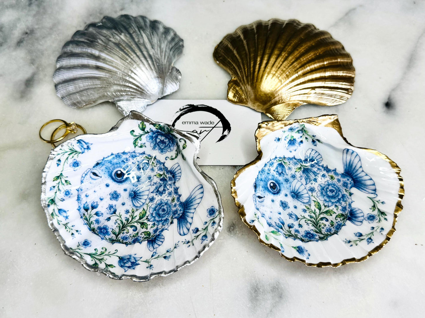 Handcrafted scallop shell trinket dish featuring an original Delft Blue puffer fish illustration created and designed by Emma Wade ART. Natural scallop shell finished with a choice of genuine gold leaf or silver leaf and sealed beneath crystal-hard glossy resin. Coastal ring holder, jewellery dish and decorative shell tray inspired by traditional Delft ceramics and marine life. Available in standard 10–11cm or large 12–14cm sizes.