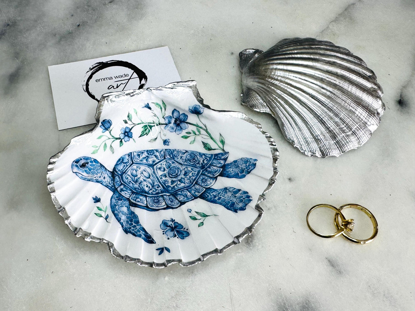 Handcrafted scallop shell trinket dish featuring an original illustrated Delft Blue Sea Turtle design by Emma Wade ART. Natural shell with hand-gilded gold or silver leaf rim and glossy resin finish. Coastal jewellery tray for rings, earrings and keepsakes. Beach house decor, sea life gift and ocean-inspired home accessory available in standard and large sizes.