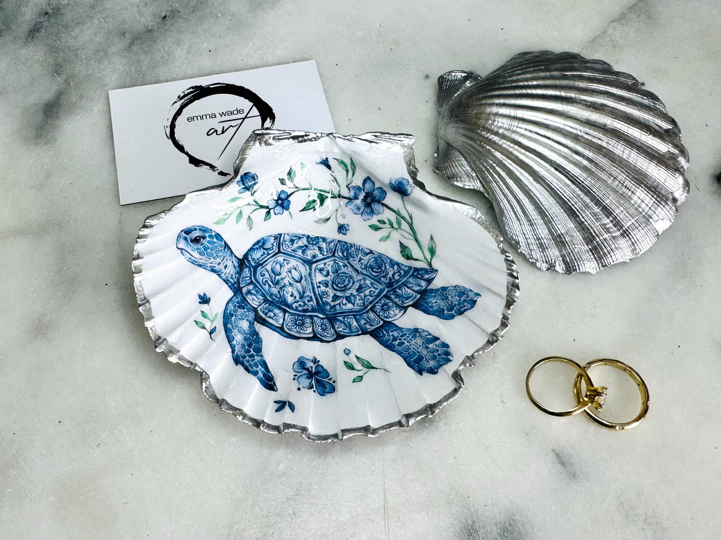 Handcrafted scallop shell trinket dish featuring an original illustrated Delft Blue Sea Turtle design by Emma Wade ART. Natural shell with hand-gilded gold or silver leaf rim and glossy resin finish. Coastal jewellery tray for rings, earrings and keepsakes. Beach house decor, sea life gift and ocean-inspired home accessory available in standard and large sizes.