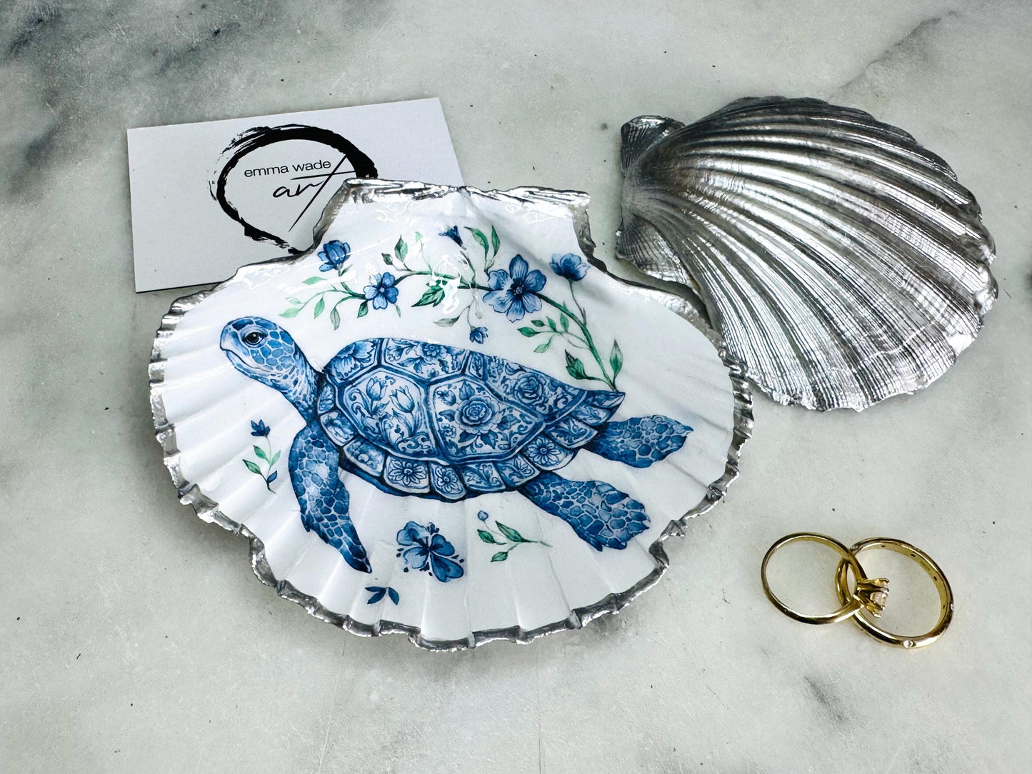 Handcrafted scallop shell trinket dish featuring an original illustrated Delft Blue Sea Turtle design by Emma Wade ART. Natural shell with hand-gilded gold or silver leaf rim and glossy resin finish. Coastal jewellery tray for rings, earrings and keepsakes. Beach house decor, sea life gift and ocean-inspired home accessory available in standard and large sizes.