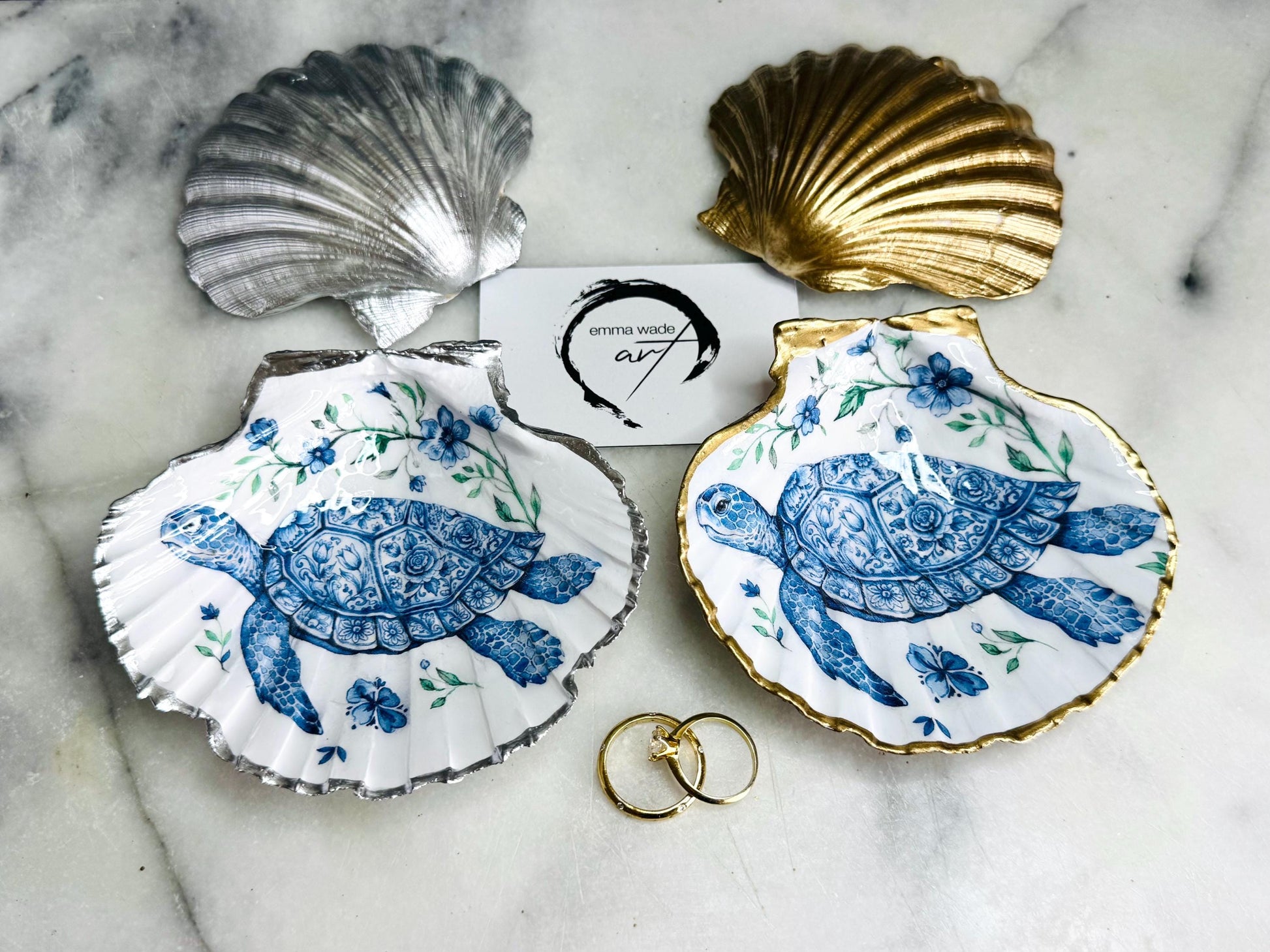 Handcrafted scallop shell trinket dish featuring an original illustrated Delft Blue Sea Turtle design by Emma Wade ART. Natural shell with hand-gilded gold or silver leaf rim and glossy resin finish. Coastal jewellery tray for rings, earrings and keepsakes. Beach house decor, sea life gift and ocean-inspired home accessory available in standard and large sizes.
