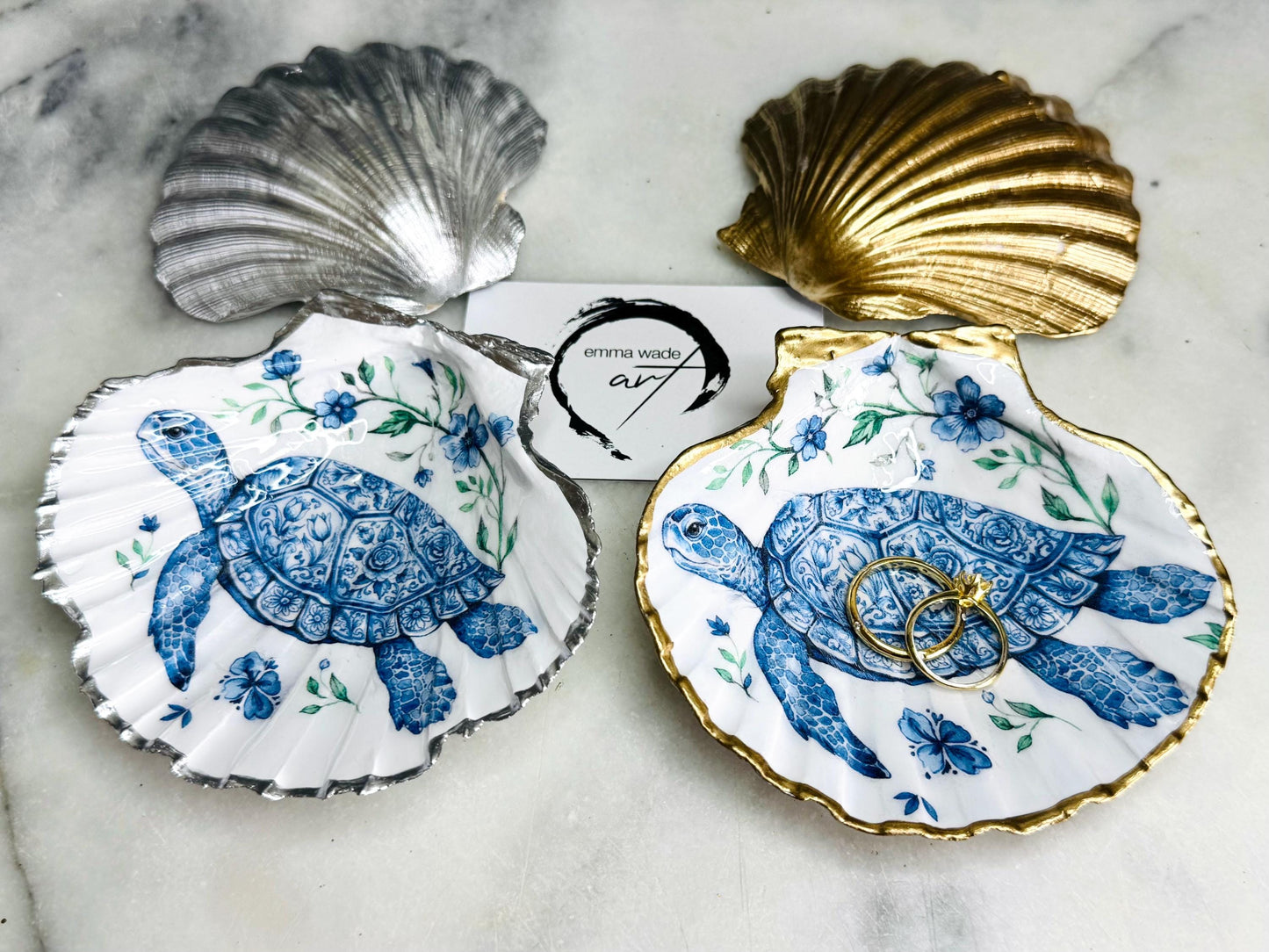 Handcrafted scallop shell trinket dish featuring an original illustrated Delft Blue Sea Turtle design by Emma Wade ART. Natural shell with hand-gilded gold or silver leaf rim and glossy resin finish. Coastal jewellery tray for rings, earrings and keepsakes. Beach house decor, sea life gift and ocean-inspired home accessory available in standard and large sizes.