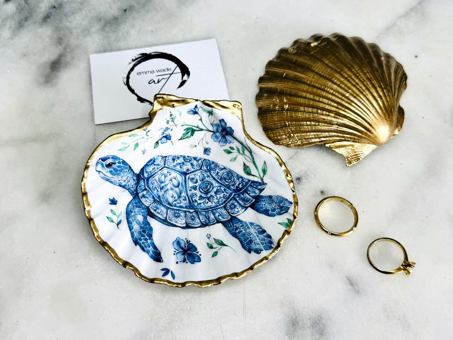 Handcrafted scallop shell trinket dish featuring an original illustrated Delft Blue Sea Turtle design by Emma Wade ART. Natural shell with hand-gilded gold or silver leaf rim and glossy resin finish. Coastal jewellery tray for rings, earrings and keepsakes. Beach house decor, sea life gift and ocean-inspired home accessory available in standard and large sizes.