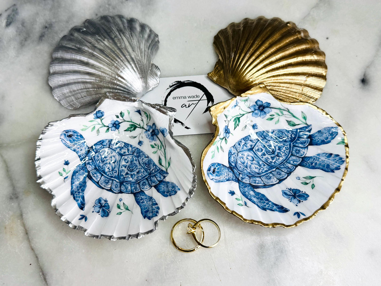 Handcrafted scallop shell trinket dish featuring an original illustrated Delft Blue Sea Turtle design by Emma Wade ART. Natural shell with hand-gilded gold or silver leaf rim and glossy resin finish. Coastal jewellery tray for rings, earrings and keepsakes. Beach house decor, sea life gift and ocean-inspired home accessory available in standard and large sizes.
