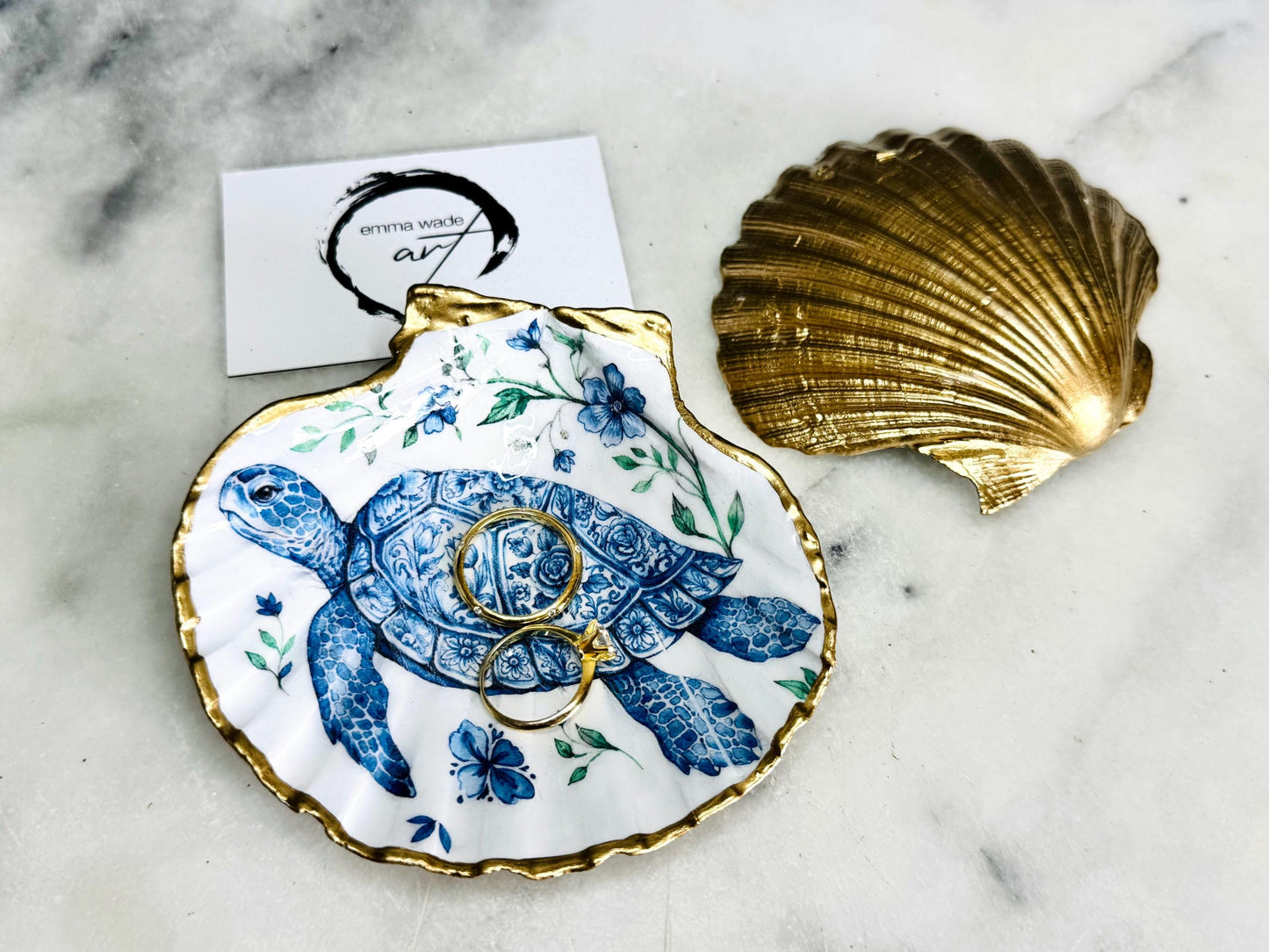 Handcrafted scallop shell trinket dish featuring an original illustrated Delft Blue Sea Turtle design by Emma Wade ART. Natural shell with hand-gilded gold or silver leaf rim and glossy resin finish. Coastal jewellery tray for rings, earrings and keepsakes. Beach house decor, sea life gift and ocean-inspired home accessory available in standard and large sizes.