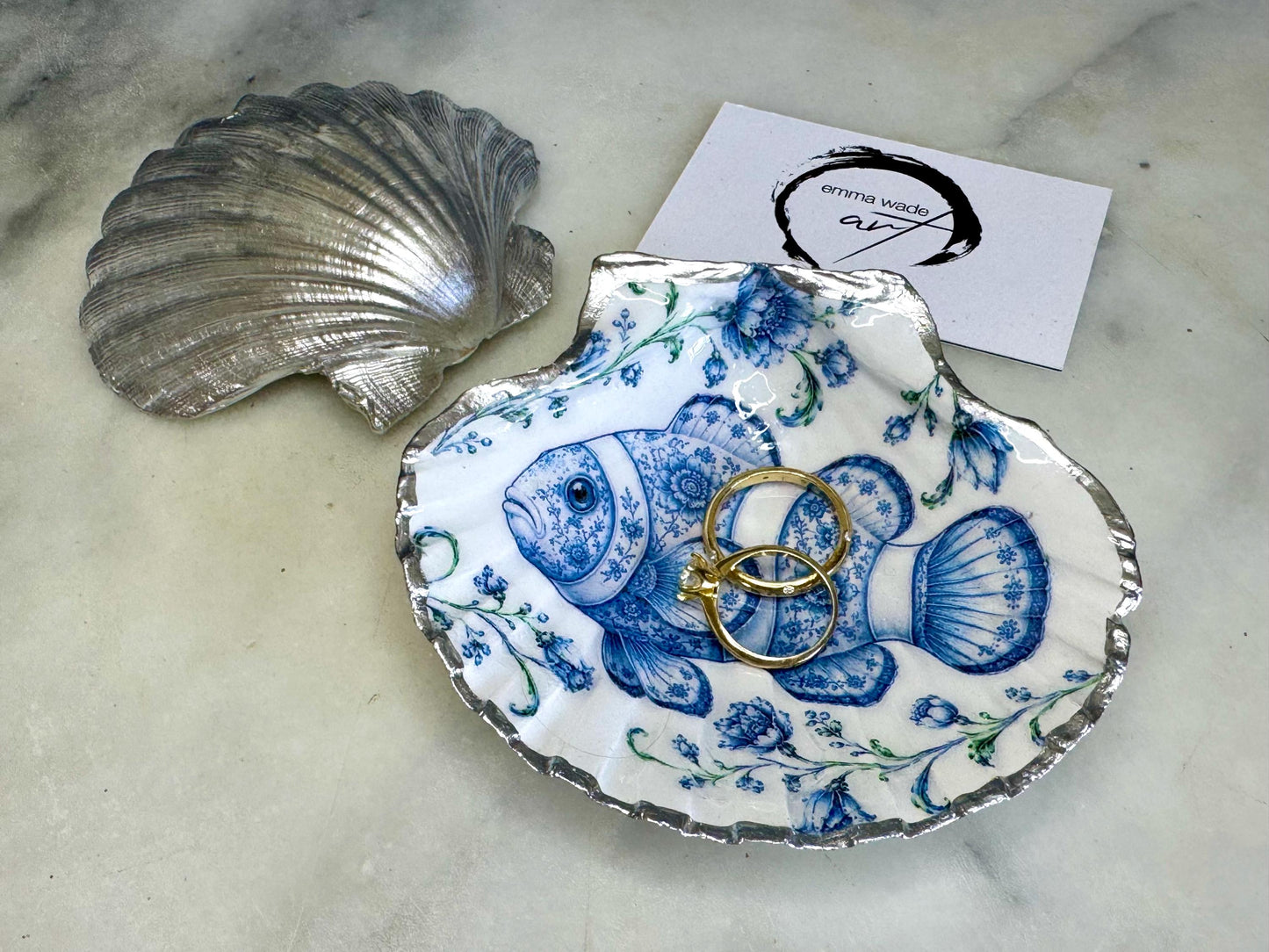 Clownfish scallop shell trinket dish featuring an original illustrated Delft Blue design by Emma Wade ART. Natural scallop shell finished with gold leaf or silver leaf edging and a crystal-clear glossy resin coating. Coastal jewellery tray, ring holder and ocean-inspired gift for fish and sea lovers, beach house décor, weddings, bridesmaids and nautical homes.