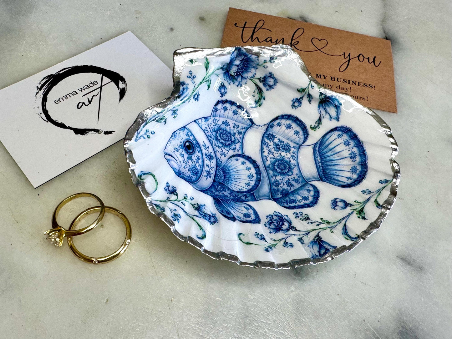 Clownfish scallop shell trinket dish featuring an original illustrated Delft Blue design by Emma Wade ART. Natural scallop shell finished with gold leaf or silver leaf edging and a crystal-clear glossy resin coating. Coastal jewellery tray, ring holder and ocean-inspired gift for fish and sea lovers, beach house décor, weddings, bridesmaids and nautical homes.