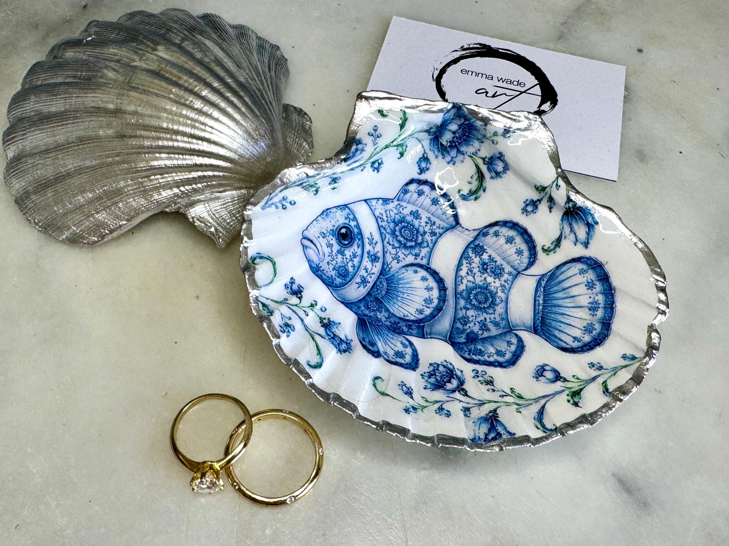 Clownfish scallop shell trinket dish featuring an original illustrated Delft Blue design by Emma Wade ART. Natural scallop shell finished with gold leaf or silver leaf edging and a crystal-clear glossy resin coating. Coastal jewellery tray, Floral ring holder and ocean-inspired gift for fish and sea lovers, beach house décor, weddings, bridesmaids and nautical homes.