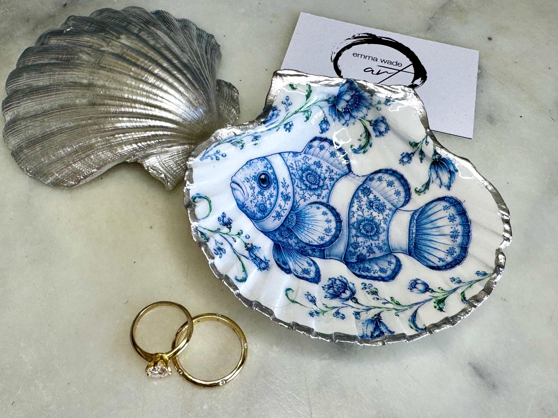 Clownfish scallop shell trinket dish featuring an original illustrated Delft Blue design by Emma Wade ART. Natural scallop shell finished with gold leaf or silver leaf edging and a crystal-clear glossy resin coating. Coastal jewellery tray, Floral ring holder and ocean-inspired gift for fish and sea lovers, beach house décor, weddings, bridesmaids and nautical homes.