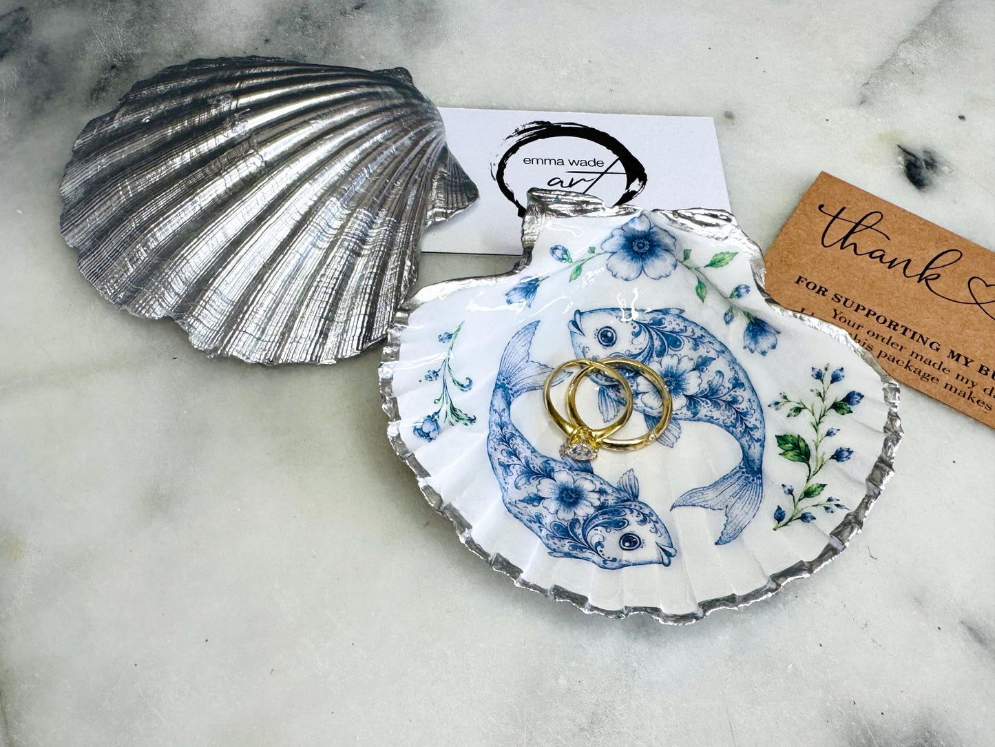 Handcrafted scallop shell trinket dish featuring an original Delft Blue fish illustration created and designed by Emma Wade ART. Natural scallop shell finished with a choice of genuine gold leaf or silver leaf and sealed beneath crystal-hard glossy resin. Coastal ring holder, jewellery dish and decorative shell tray inspired by traditional Delft ceramics and marine life. Available in standard 10–11cm or large 12–14cm sizes. Part of the Delft Blue Sea Life Collection.