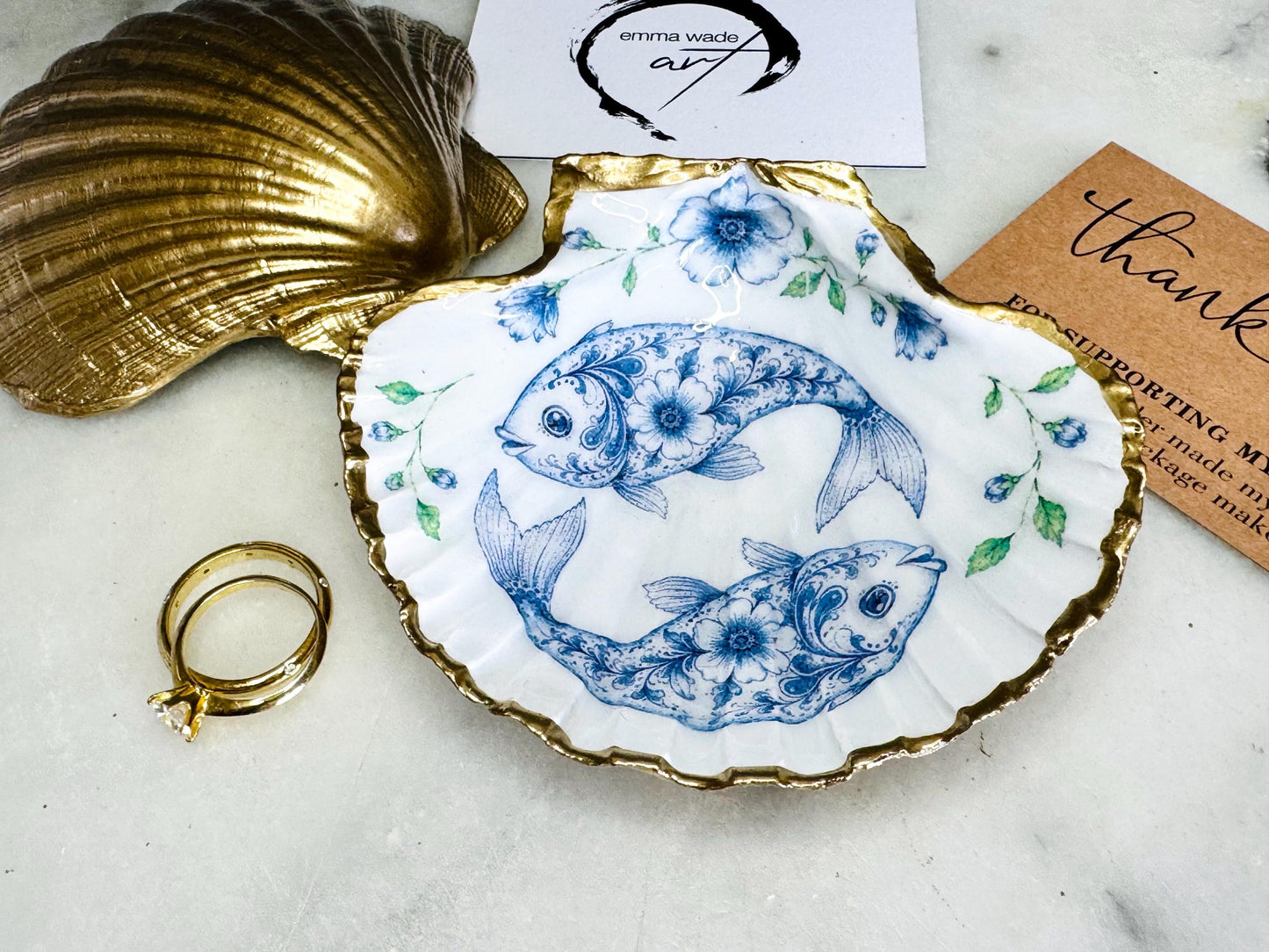 Handcrafted scallop shell trinket dish featuring an original Delft Blue fish illustration created and designed by Emma Wade ART. Natural scallop shell finished with a choice of genuine gold leaf or silver leaf and sealed beneath crystal-hard glossy resin. Coastal ring holder, jewellery dish and decorative shell tray inspired by traditional Delft ceramics and marine life. Available in standard 10–11cm or large 12–14cm sizes. Part of the Delft Blue Sea Life Collection.