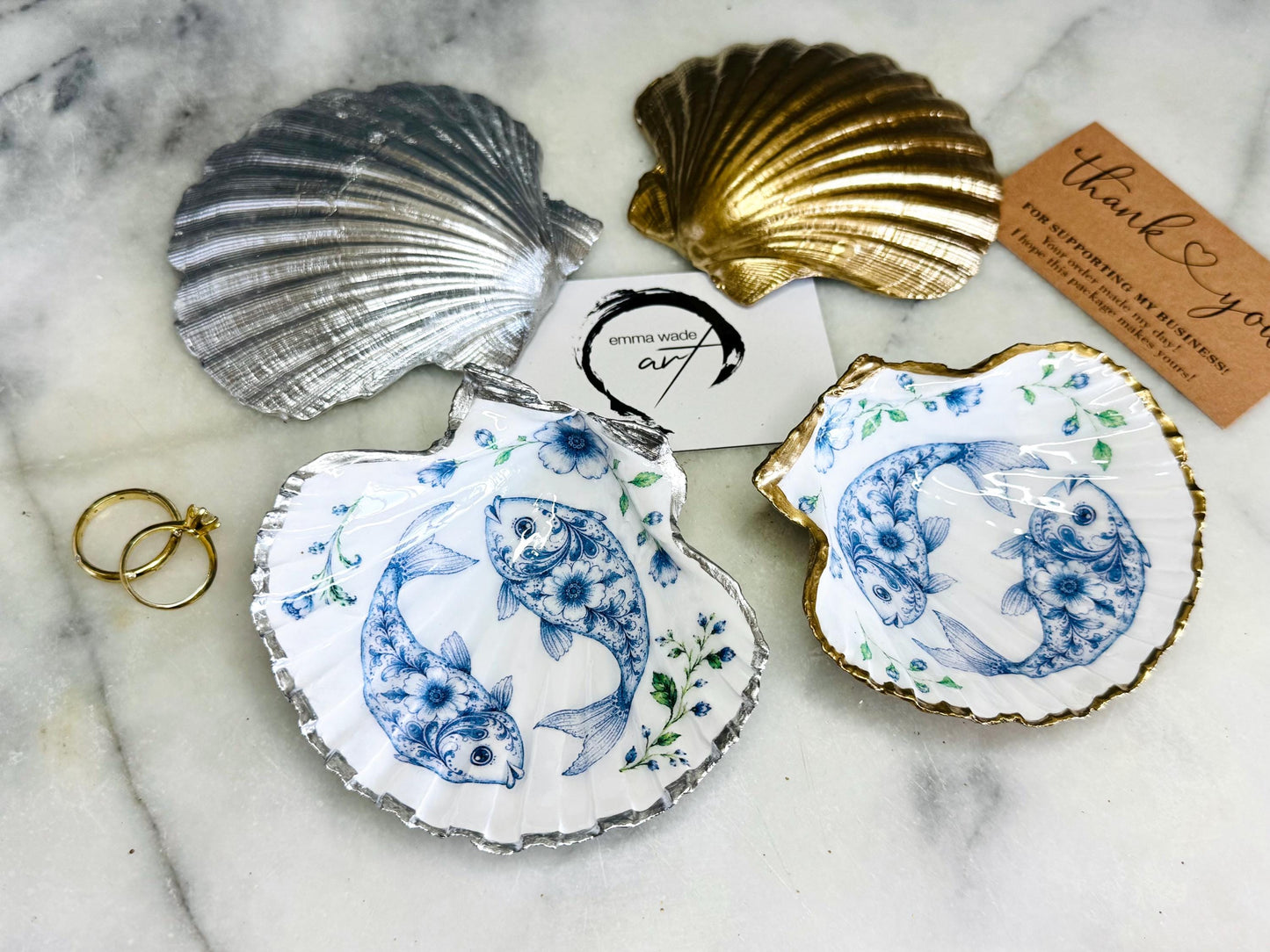 Handcrafted scallop shell trinket dish featuring an original Delft Blue fish illustration created and designed by Emma Wade ART. Natural scallop shell finished with a choice of genuine gold leaf or silver leaf and sealed beneath crystal-hard glossy resin. Coastal ring holder, jewellery dish and decorative shell tray inspired by traditional Delft ceramics and marine life. Available in standard 10–11cm or large 12–14cm sizes. Part of the Delft Blue Sea Life Collection.