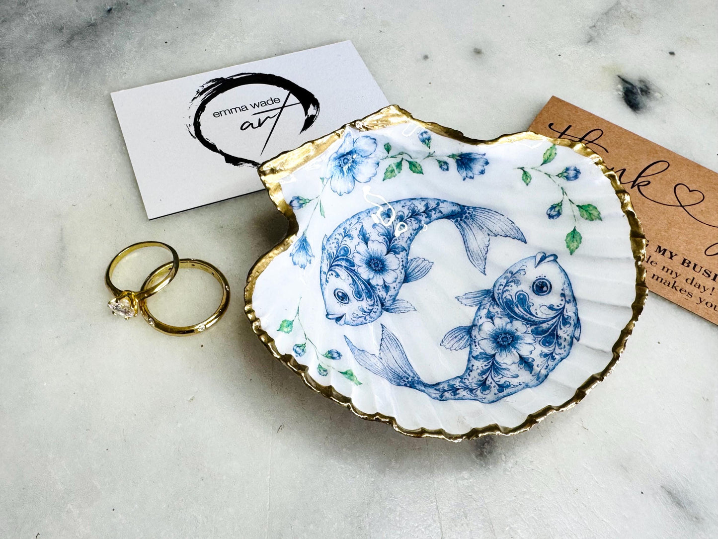 Handcrafted scallop shell trinket dish featuring an original Delft Blue fish illustration created and designed by Emma Wade ART. Natural scallop shell finished with a choice of genuine gold leaf or silver leaf and sealed beneath crystal-hard glossy resin. Coastal ring holder, jewellery dish and decorative shell tray inspired by traditional Delft ceramics and marine life. Available in standard 10–11cm or large 12–14cm sizes. Part of the Delft Blue Sea Life Collection.