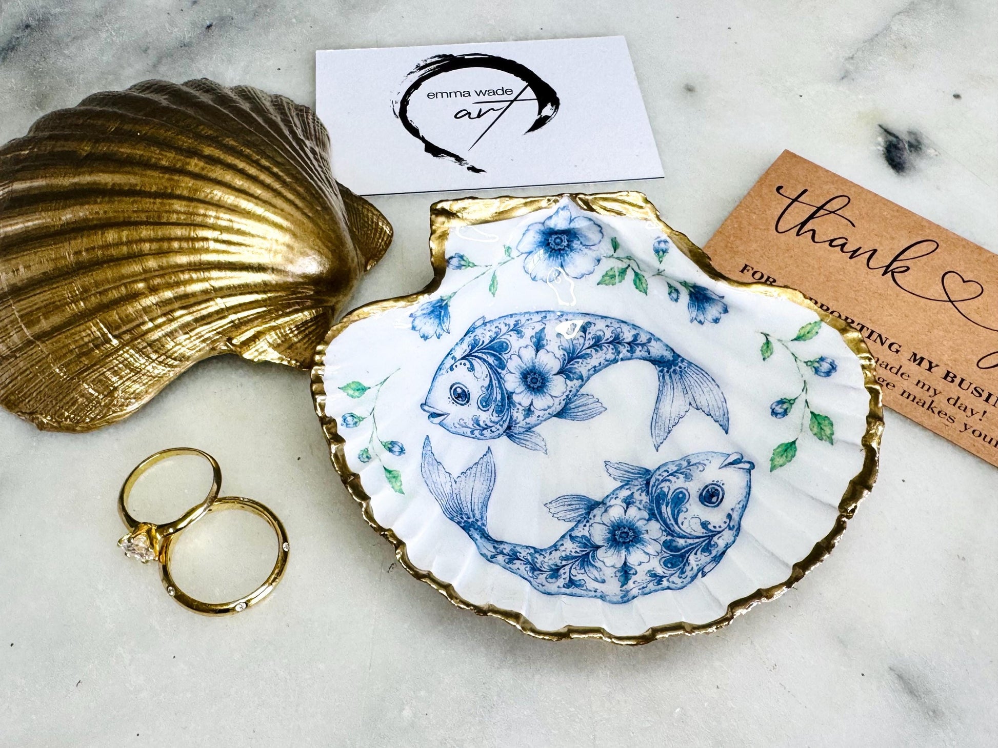 Handcrafted scallop shell trinket dish featuring an original Delft Blue fish illustration created and designed by Emma Wade ART. Natural scallop shell finished with a choice of genuine gold leaf or silver leaf and sealed beneath crystal-hard glossy resin. Coastal ring holder, jewellery dish and decorative shell tray inspired by traditional Delft ceramics and marine life. Available in standard 10–11cm or large 12–14cm sizes. Part of the Delft Blue Sea Life Collection.