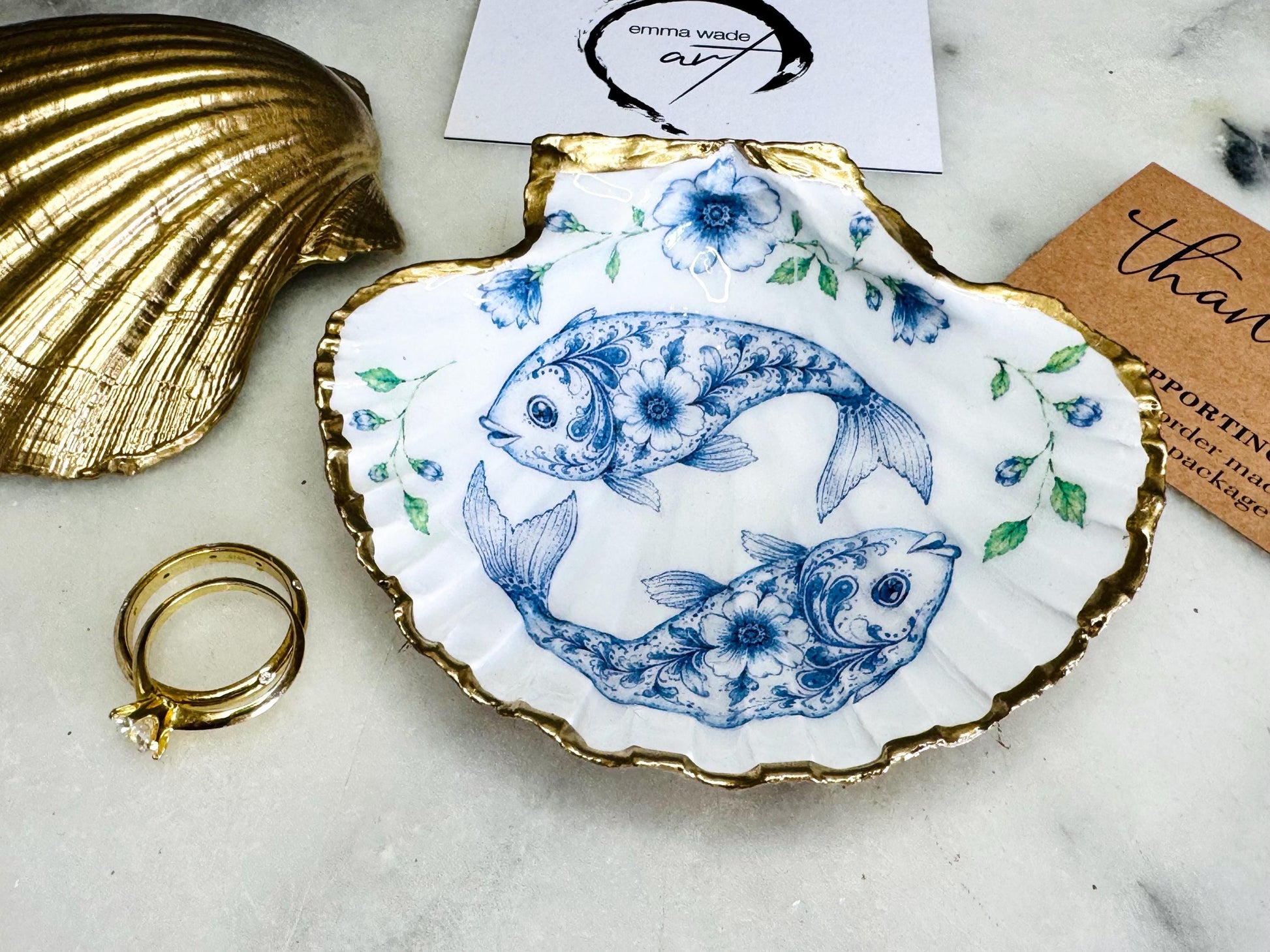 Handcrafted scallop shell trinket dish featuring an original Delft Blue fish illustration created and designed by Emma Wade ART. Natural scallop shell finished with a choice of genuine gold leaf or silver leaf and sealed beneath crystal-hard glossy resin. Coastal ring holder, jewellery dish and decorative shell tray inspired by traditional Delft ceramics and marine life. Available in standard 10–11cm or large 12–14cm sizes. Part of the Delft Blue Sea Life Collection.
