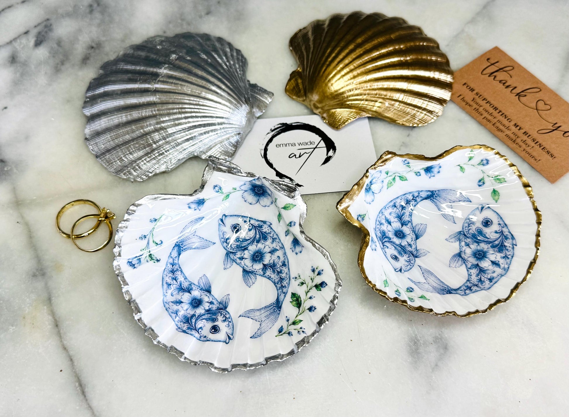 Handcrafted scallop shell trinket dish featuring an original Delft Blue fish illustration created and designed by Emma Wade ART. Natural scallop shell finished with a choice of genuine gold leaf or silver leaf and sealed beneath crystal-hard glossy resin. Coastal ring holder, jewellery dish and decorative shell tray inspired by traditional Delft ceramics and marine life. Available in standard 10–11cm or large 12–14cm sizes. Part of the Delft Blue Sea Life Collection.