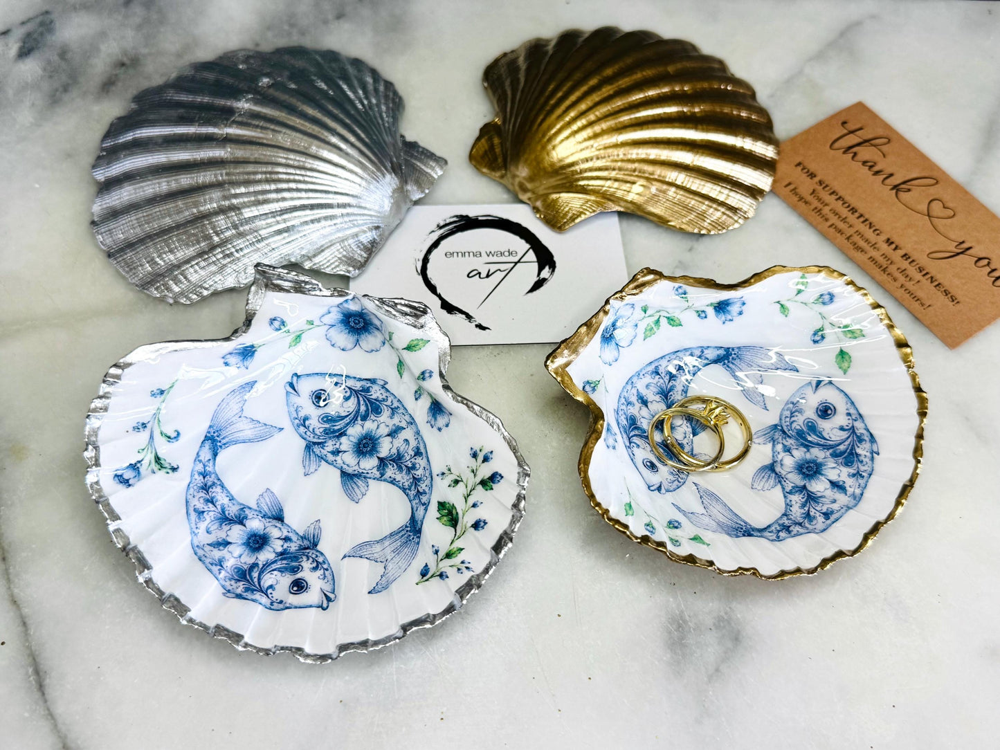 Handcrafted scallop shell trinket dish featuring an original Delft Blue fish illustration created and designed by Emma Wade ART. Natural scallop shell finished with a choice of genuine gold leaf or silver leaf and sealed beneath crystal-hard glossy resin. Coastal ring holder, jewellery dish and decorative shell tray inspired by traditional Delft ceramics and marine life. Available in standard 10–11cm or large 12–14cm sizes. Part of the Delft Blue Sea Life Collection.