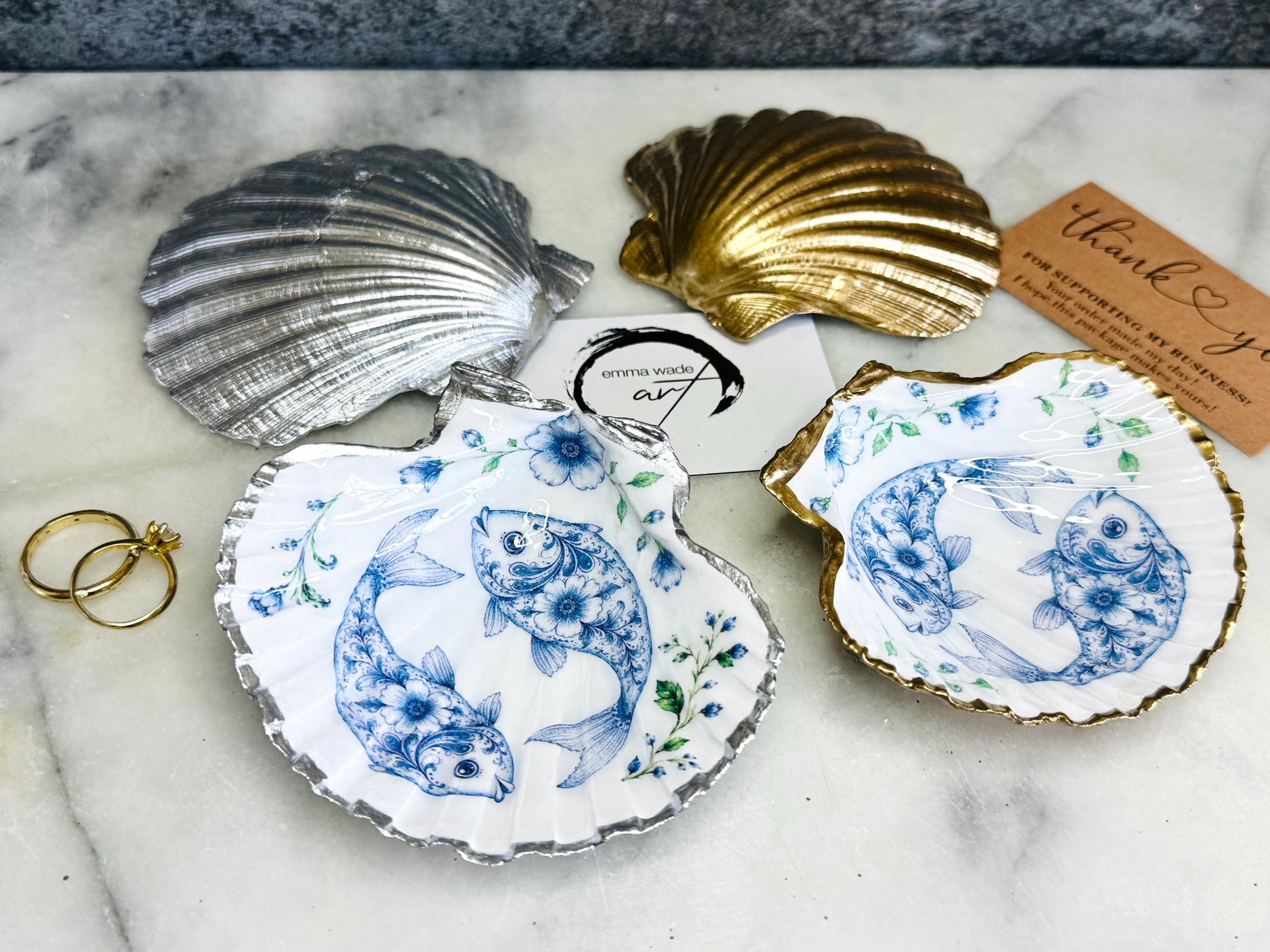 Handcrafted scallop shell trinket dish featuring an original Delft Blue fish illustration created and designed by Emma Wade ART. Natural scallop shell finished with a choice of genuine gold leaf or silver leaf and sealed beneath crystal-hard glossy resin. Coastal ring holder, jewellery dish and decorative shell tray inspired by traditional Delft ceramics and marine life. Available in standard 10–11cm or large 12–14cm sizes. Part of the Delft Blue Sea Life Collection.