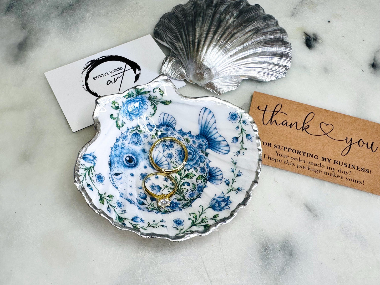 Handcrafted scallop shell trinket dish featuring an original Delft Blue puffer fish illustration created and designed by Emma Wade ART. Natural scallop shell finished with a choice of genuine gold leaf or silver leaf and sealed beneath crystal-hard glossy resin. Coastal ring holder, jewellery dish and decorative shell tray inspired by traditional Delft ceramics and marine life. Available in standard 10–11cm or large 12–14cm sizes.