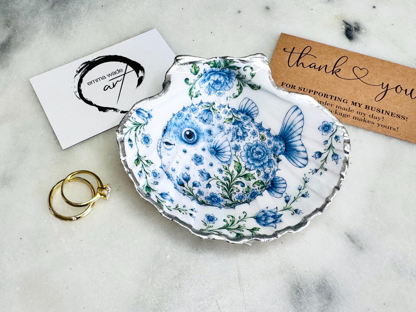 Handcrafted scallop shell trinket dish featuring an original Delft Blue puffer fish illustration created and designed by Emma Wade ART. Natural scallop shell finished with a choice of genuine gold leaf or silver leaf and sealed beneath crystal-hard glossy resin. Coastal ring holder, jewellery dish and decorative shell tray inspired by traditional Delft ceramics and marine life. Available in standard 10–11cm or large 12–14cm sizes.