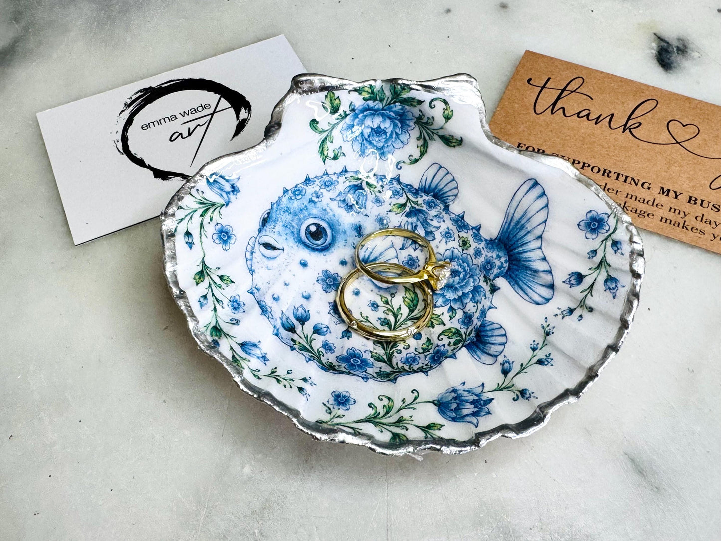 Handcrafted scallop shell trinket dish featuring an original Delft Blue puffer fish illustration created and designed by Emma Wade ART. Natural scallop shell finished with a choice of genuine gold leaf or silver leaf and sealed beneath crystal-hard glossy resin. Coastal ring holder, jewellery dish and decorative shell tray inspired by traditional Delft ceramics and marine life. Available in standard 10–11cm or large 12–14cm sizes.