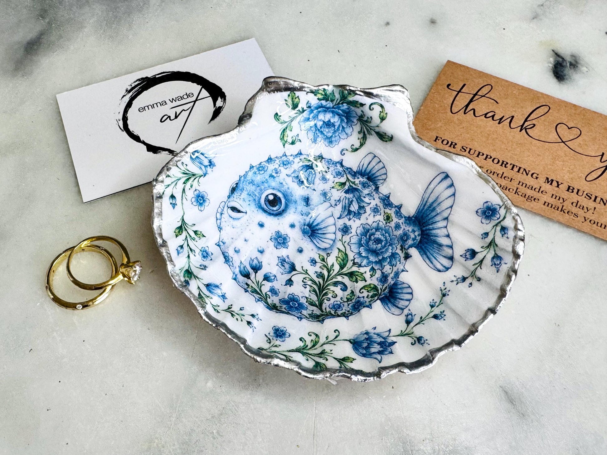 Handcrafted scallop shell trinket dish featuring an original Delft Blue puffer fish illustration created and designed by Emma Wade ART. Natural scallop shell finished with a choice of genuine gold leaf or silver leaf and sealed beneath crystal-hard glossy resin. Coastal ring holder, jewellery dish and decorative shell tray inspired by traditional Delft ceramics and marine life. Available in standard 10–11cm or large 12–14cm sizes.