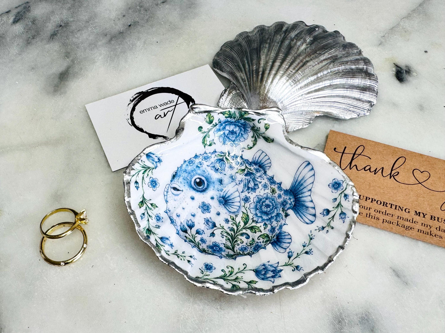 Handcrafted scallop shell trinket dish featuring an original Delft Blue puffer fish illustration created and designed by Emma Wade ART. Natural scallop shell finished with a choice of genuine gold leaf or silver leaf and sealed beneath crystal-hard glossy resin. Coastal ring holder, jewellery dish and decorative shell tray inspired by traditional Delft ceramics and marine life. Available in standard 10–11cm or large 12–14cm sizes.