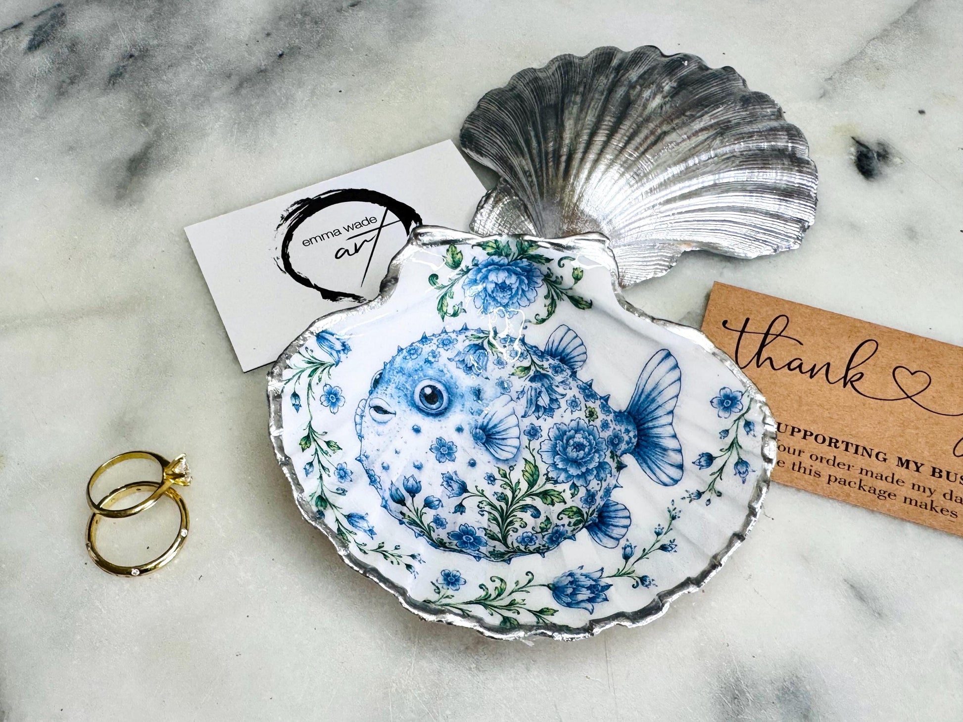 Handcrafted scallop shell trinket dish featuring an original Delft Blue puffer fish illustration created and designed by Emma Wade ART. Natural scallop shell finished with a choice of genuine gold leaf or silver leaf and sealed beneath crystal-hard glossy resin. Coastal ring holder, jewellery dish and decorative shell tray inspired by traditional Delft ceramics and marine life. Available in standard 10–11cm or large 12–14cm sizes.