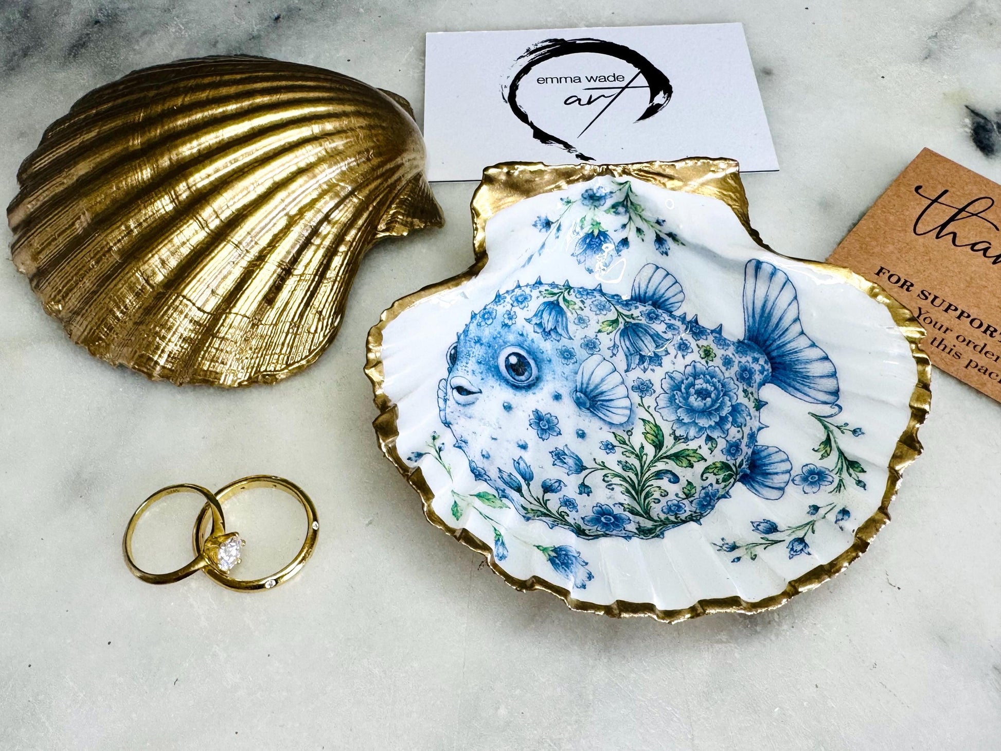 Handcrafted scallop shell trinket dish featuring an original Delft Blue puffer fish illustration created and designed by Emma Wade ART. Natural scallop shell finished with a choice of genuine gold leaf or silver leaf and sealed beneath crystal-hard glossy resin. Coastal ring holder, jewellery dish and decorative shell tray inspired by traditional Delft ceramics and marine life. Available in standard 10–11cm or large 12–14cm sizes.