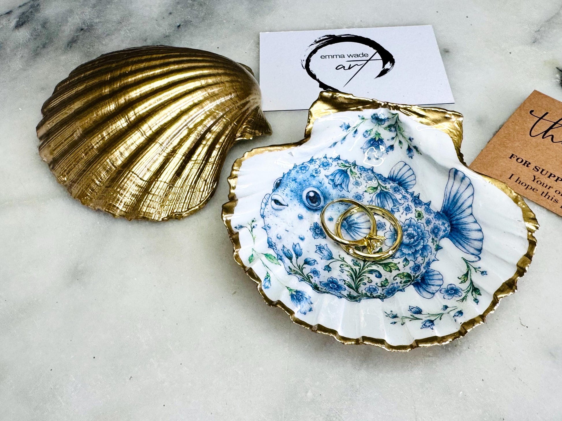 Handcrafted scallop shell trinket dish featuring an original Delft Blue puffer fish illustration created and designed by Emma Wade ART. Natural scallop shell finished with a choice of genuine gold leaf or silver leaf and sealed beneath crystal-hard glossy resin. Coastal ring holder, jewellery dish and decorative shell tray inspired by traditional Delft ceramics and marine life. Available in standard 10–11cm or large 12–14cm sizes.