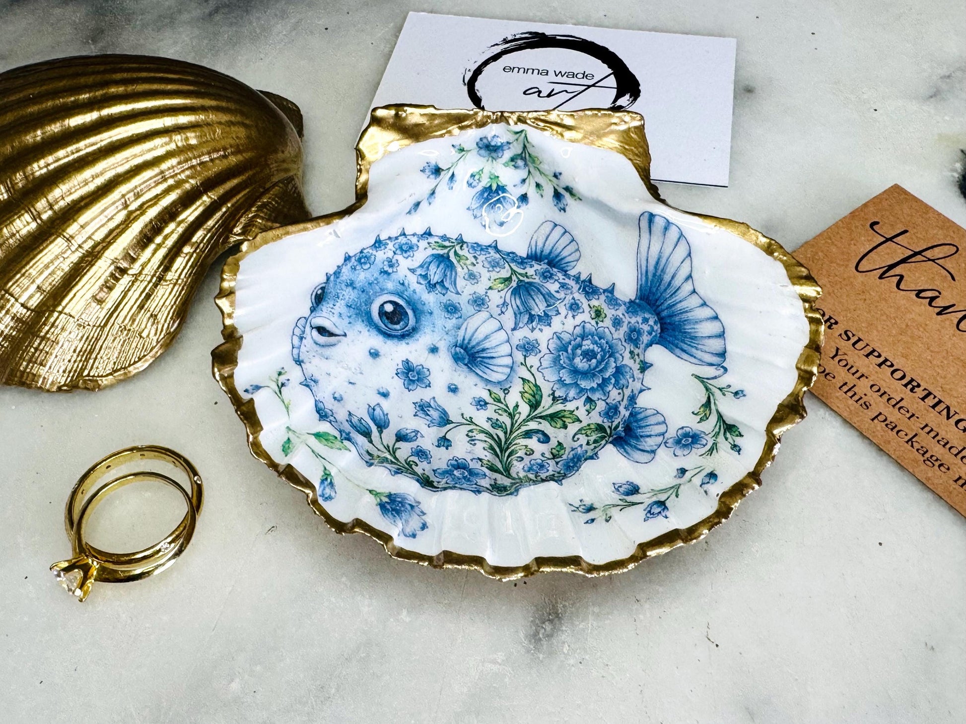 Handcrafted scallop shell trinket dish featuring an original Delft Blue puffer fish illustration created and designed by Emma Wade ART. Natural scallop shell finished with a choice of genuine gold leaf or silver leaf and sealed beneath crystal-hard glossy resin. Coastal ring holder, jewellery dish and decorative shell tray inspired by traditional Delft ceramics and marine life. Available in standard 10–11cm or large 12–14cm sizes.